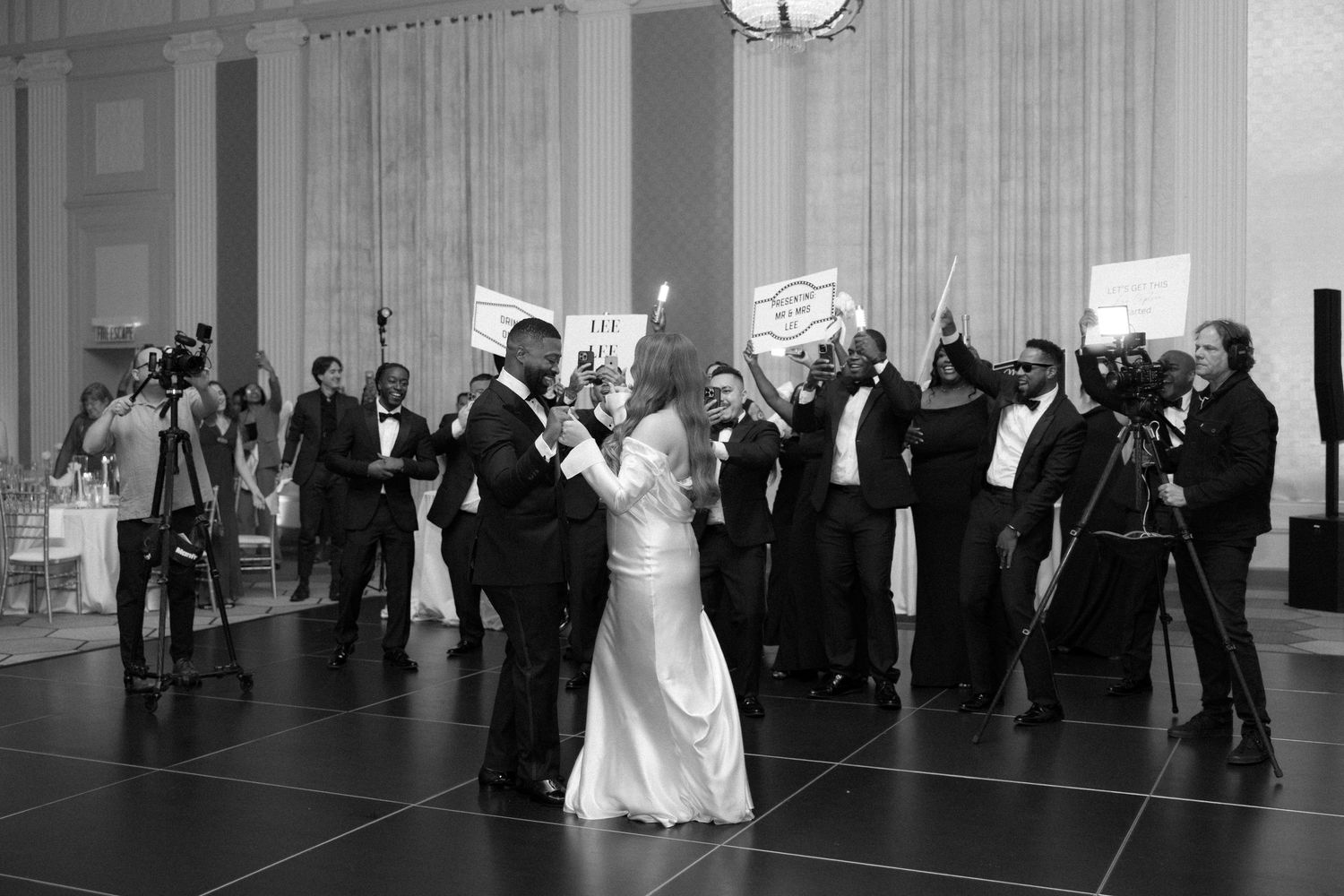 Black and white photo of a couple sharing their first dance surrounded by guests at an elegant wedding reception.