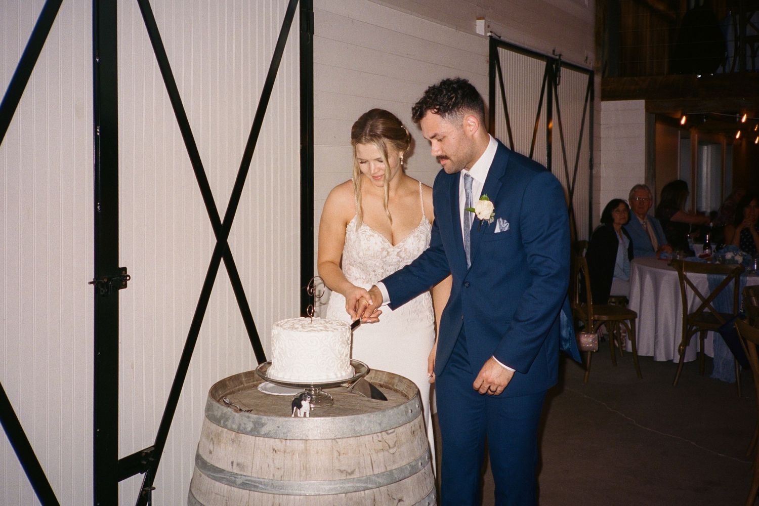 Bride and groom cutting white wedding cake on a wooden barrel at rustic indoor reception venue.