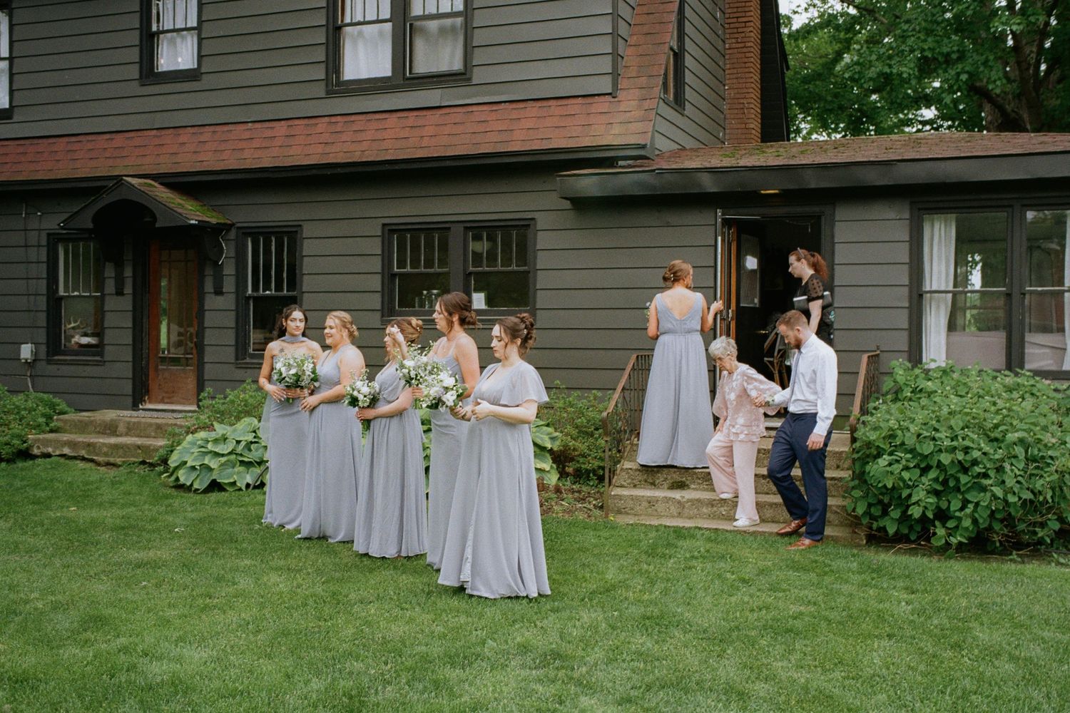 Bridesmaids in gray dresses gather outside a dark green farmhouse, holding bouquets on a lush lawn.