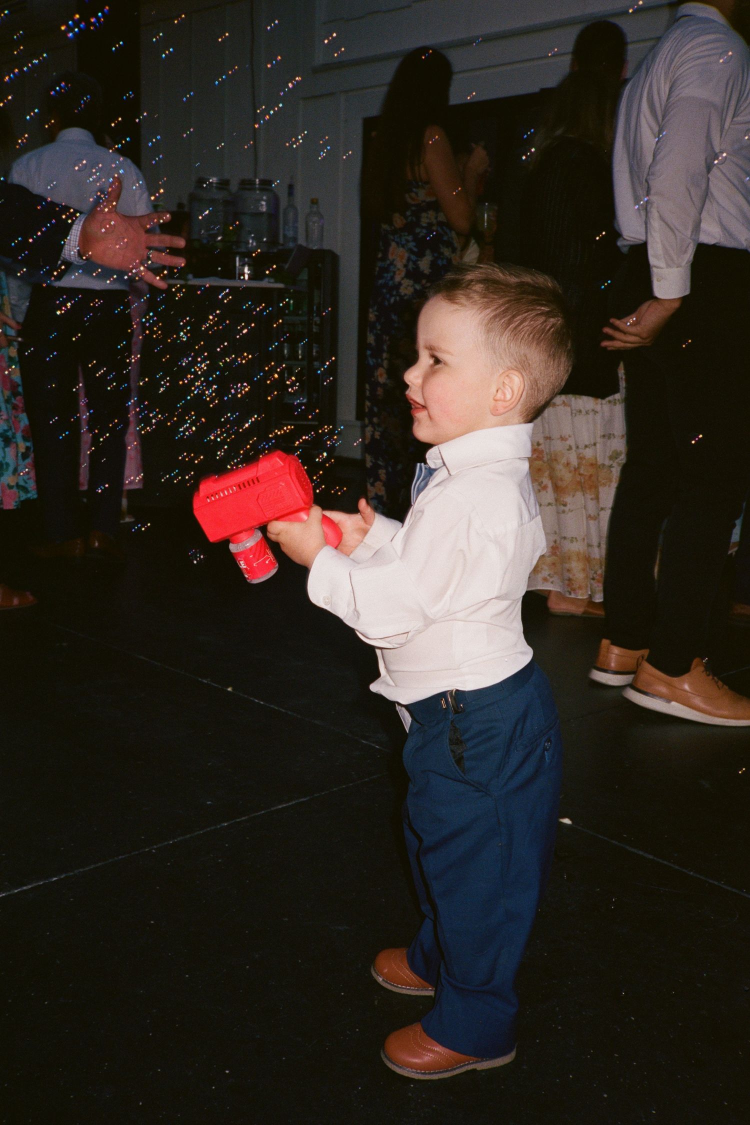 Young toddler boy in white shirt and blue pants holds red bubble gun at a festive indoor party event.