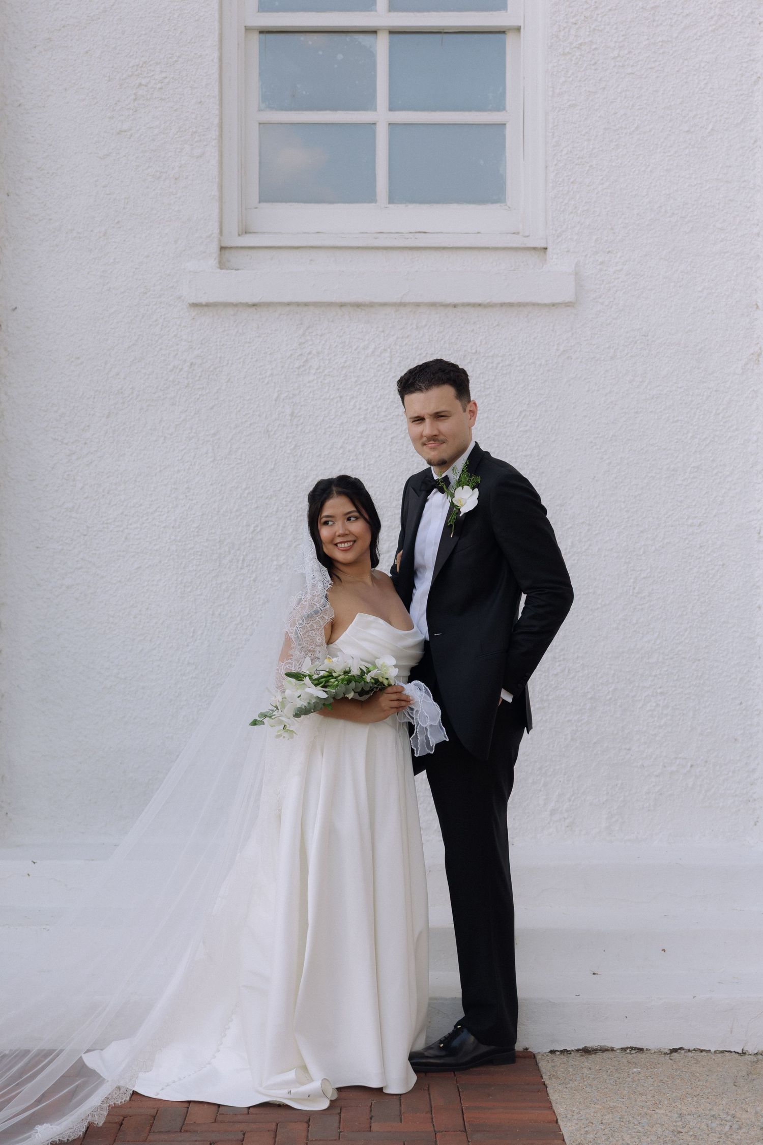 Bride and groom pose together in front of white building wall on wedding day.