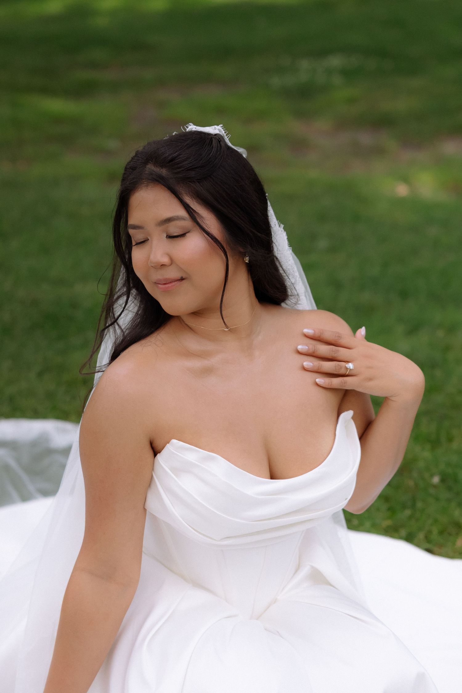 Beautiful Asian bride in strapless white gown sits on green grass with eyes closed.