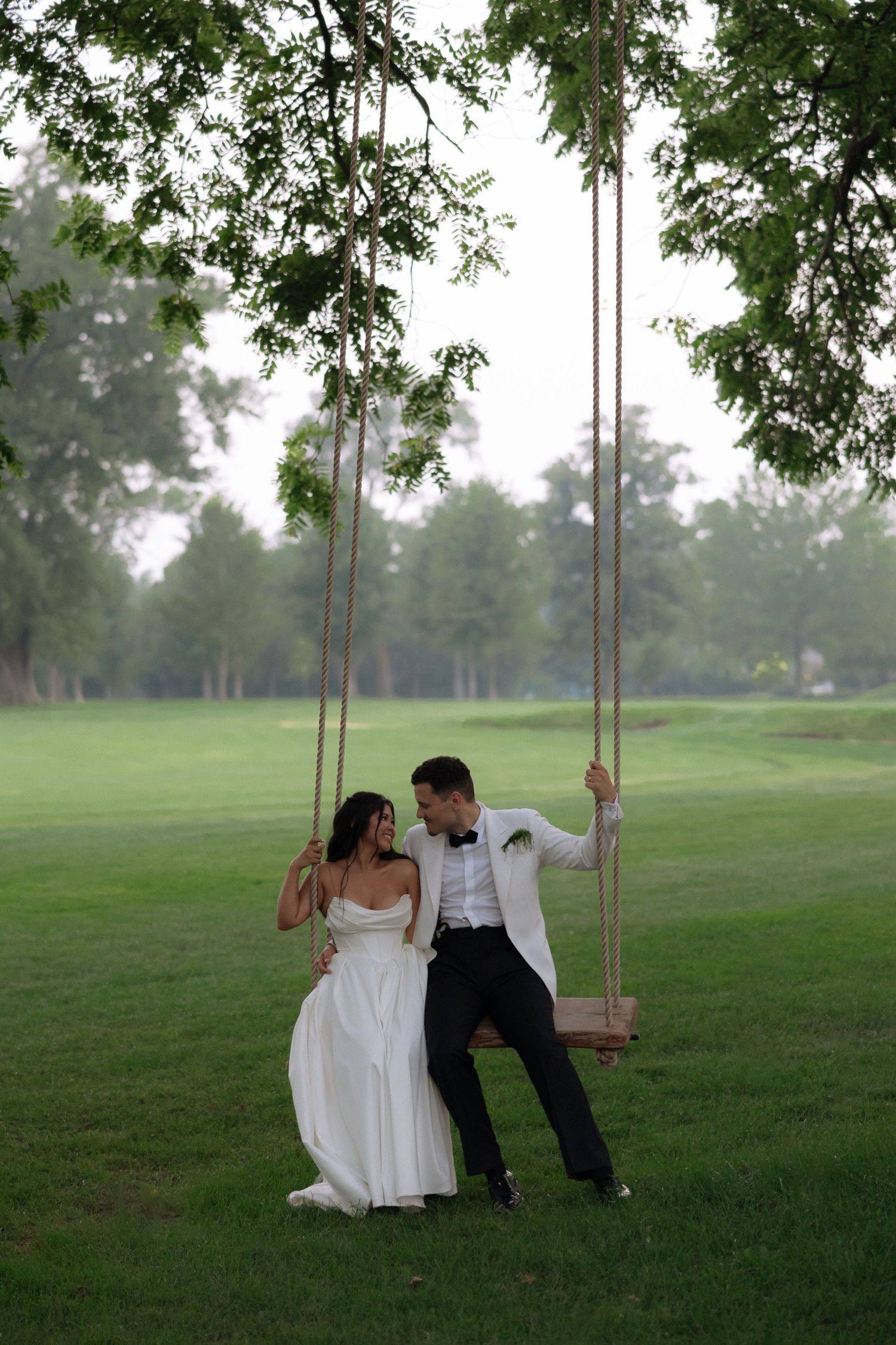 Bride in white gown stands beside groom in white tuxedo on a wooden rope swing under lush green trees.