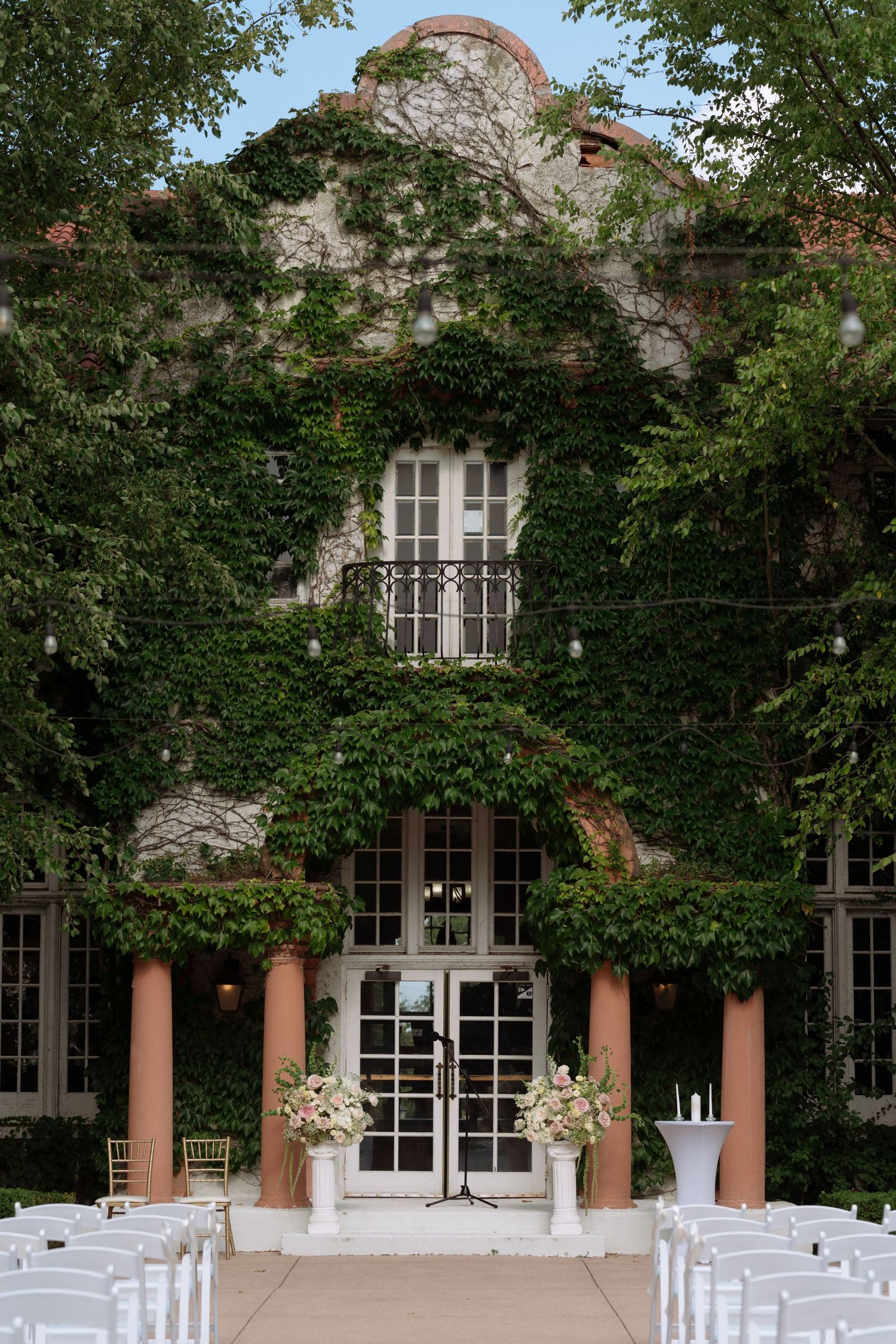 Ivy-covered stone wedding venue with white chairs, floral arrangements, and French doors set for an outdoor ceremony.