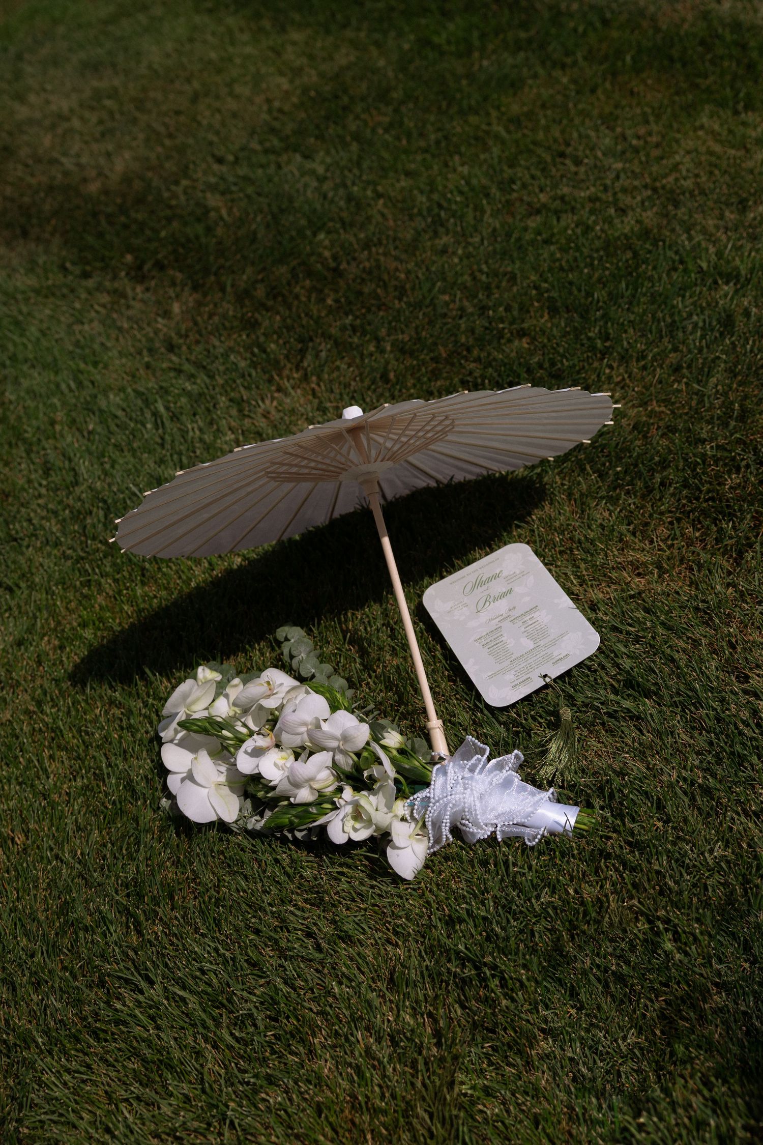 A paper parasol, white floral bouquet, and wedding program arranged on lush green grass for an outdoor ceremony.