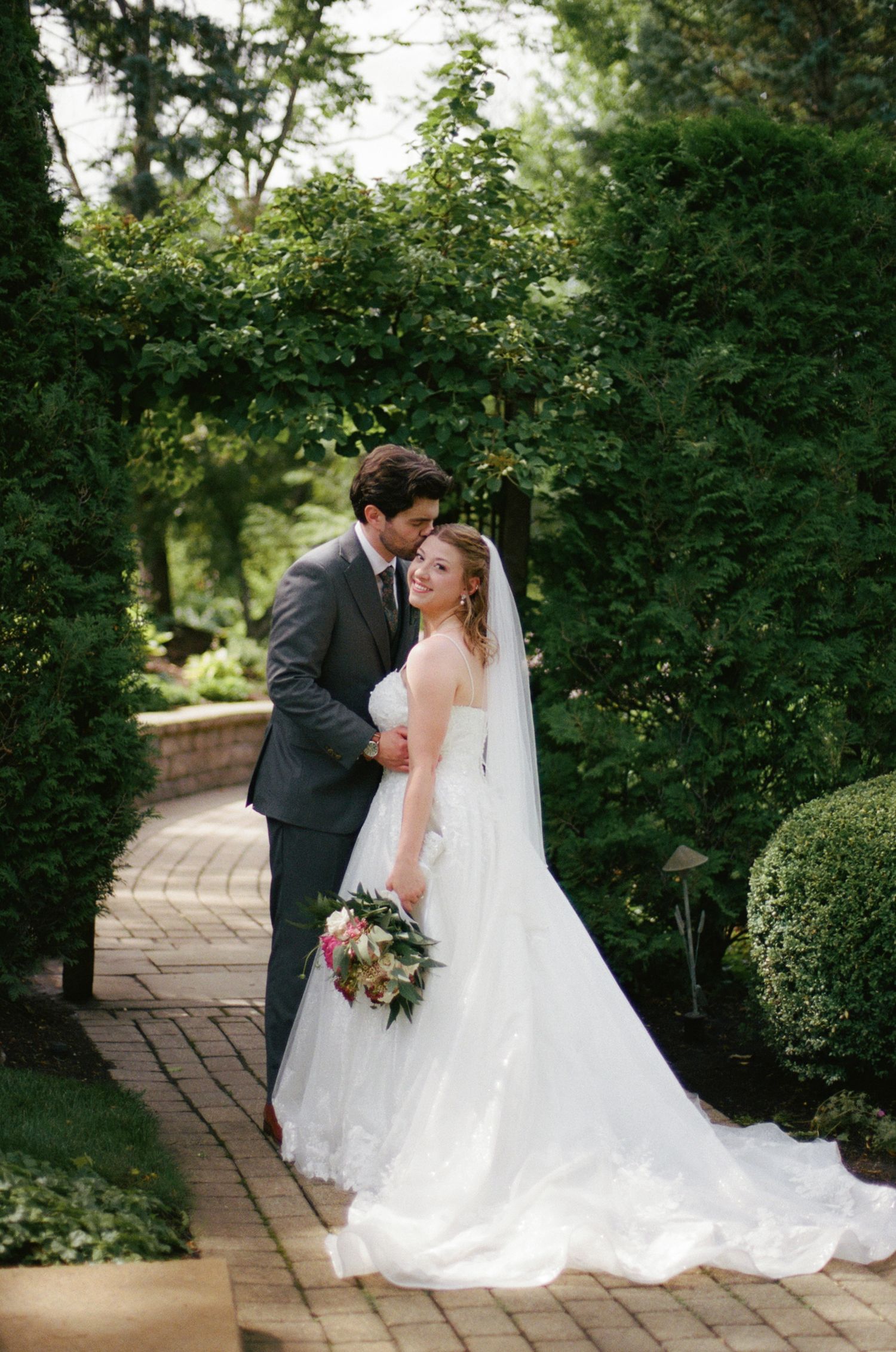 Bride and groom embrace on a brick garden path surrounded by lush green hedges, bride holding a colorful bouquet.