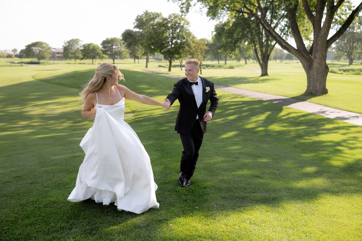 Bride and groom holding hands running across lush green golf course grounds on their wedding day.