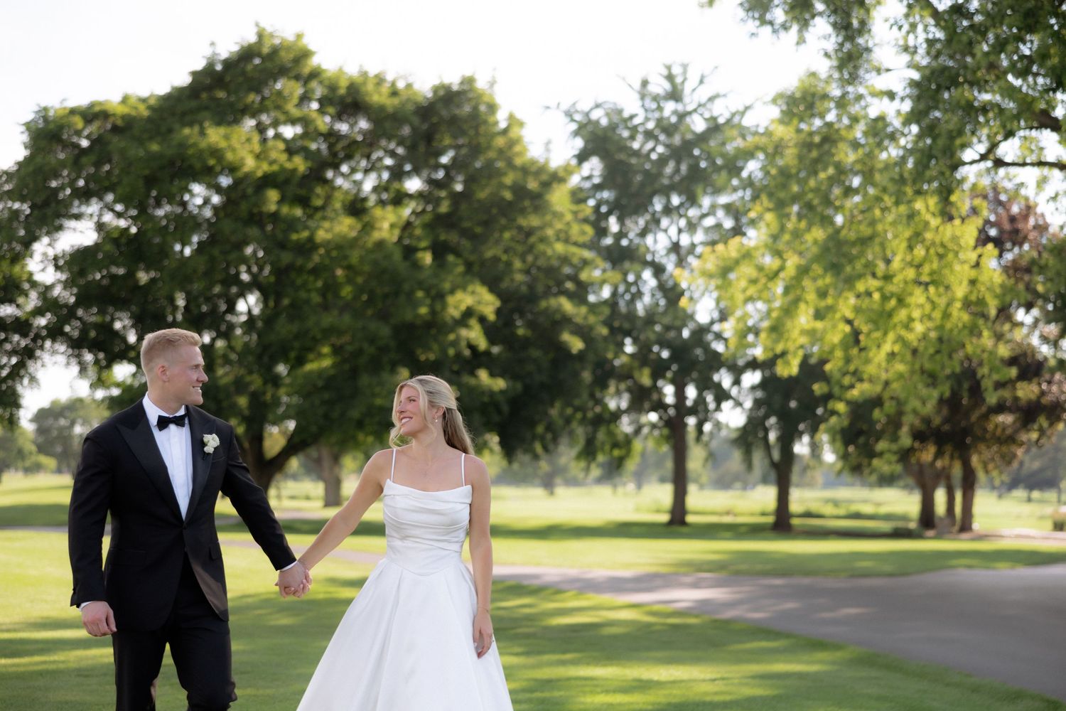 Bride and groom walk hand in hand through a scenic park, smiling joyfully on their wedding day.