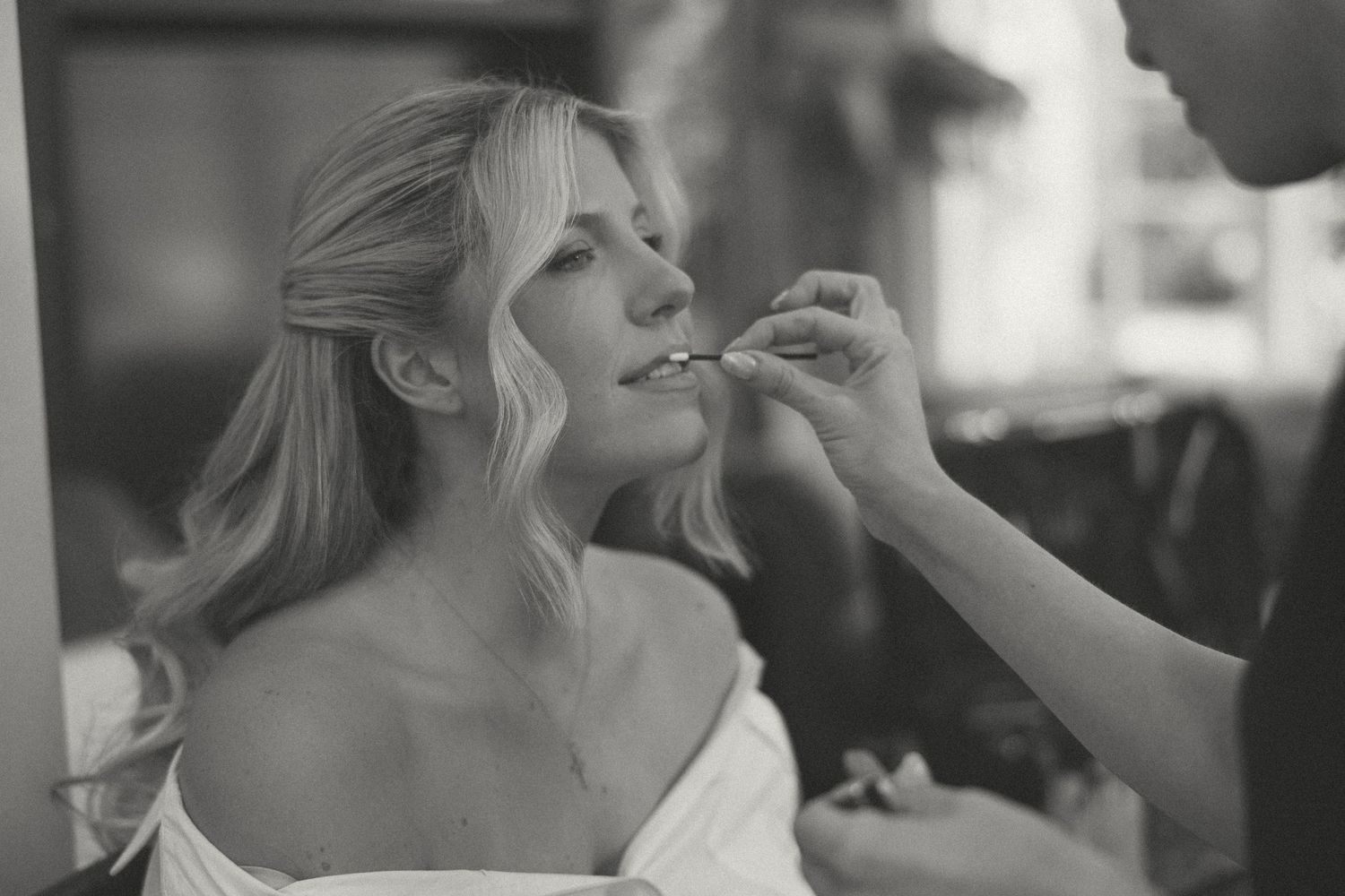 Black and white photo of a bride getting lipstick applied by a makeup artist during wedding preparations.