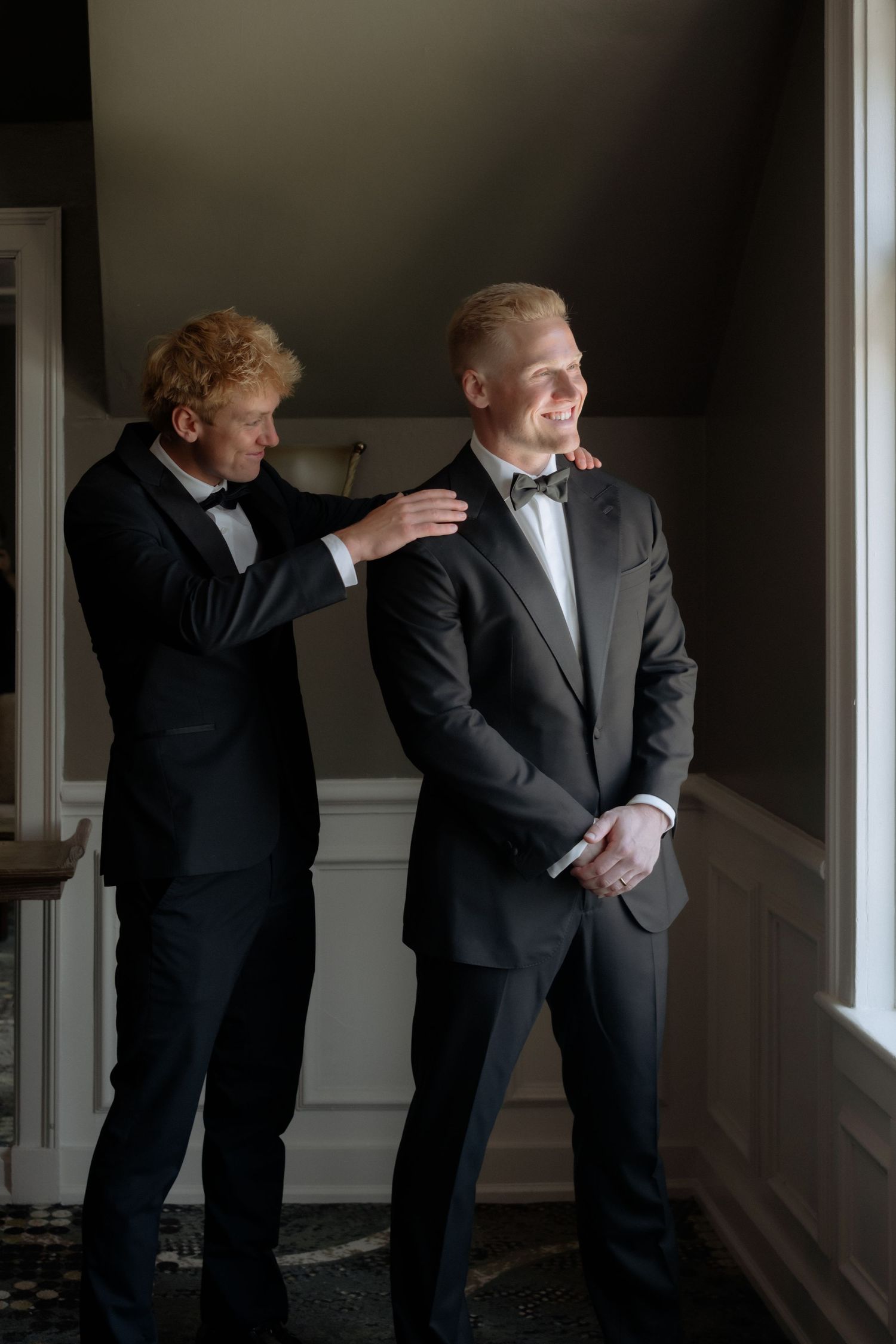 Groomsman in tuxedo helping groom adjust his black suit jacket before wedding ceremony indoors.