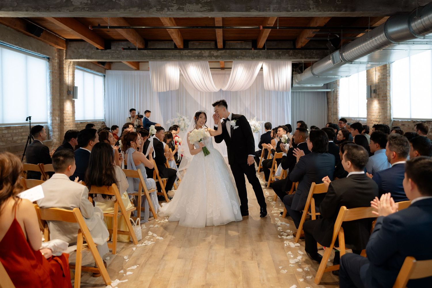 Bride and groom share a kiss at the altar during an elegant indoor wedding ceremony with guests seated in wooden chairs.