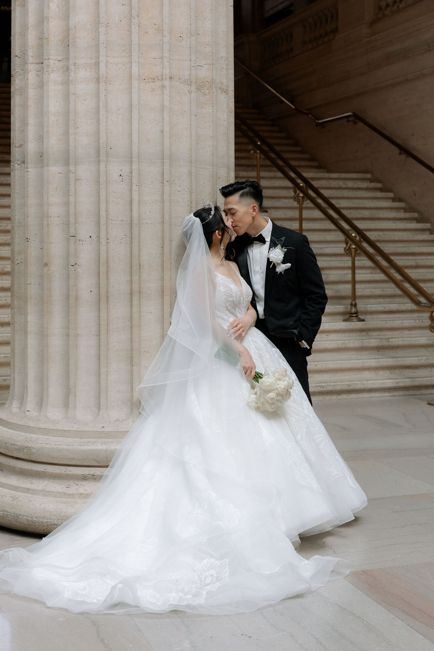 Bride and groom share romantic kiss beside grand marble columns in elegant wedding venue.