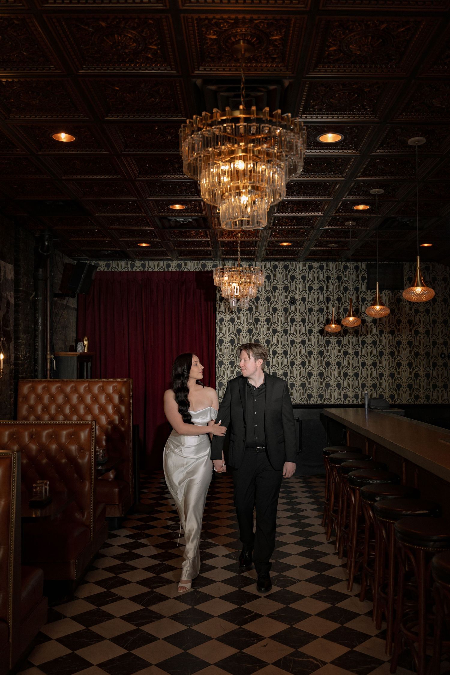 Elegant couple walks hand-in-hand beneath a crystal chandelier in a moody, vintage-styled venue with checkered floors.