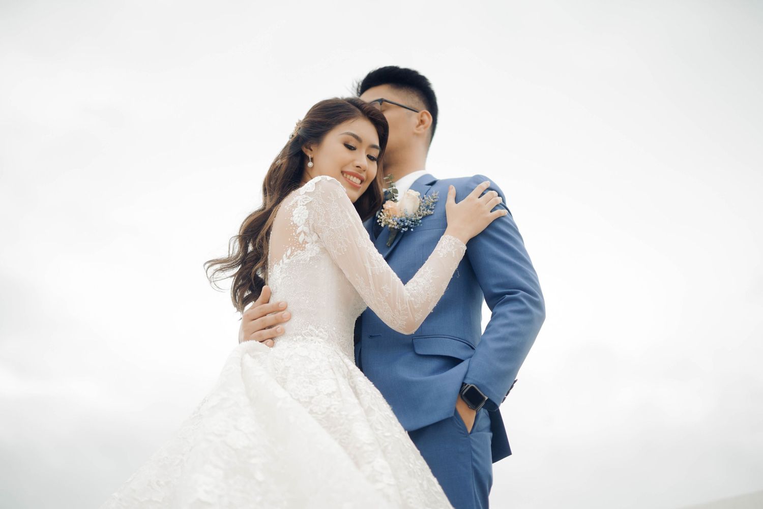 A couple in wedding attire embraces outdoors, bride in white lace gown and groom in blue suit, sharing a romantic moment.