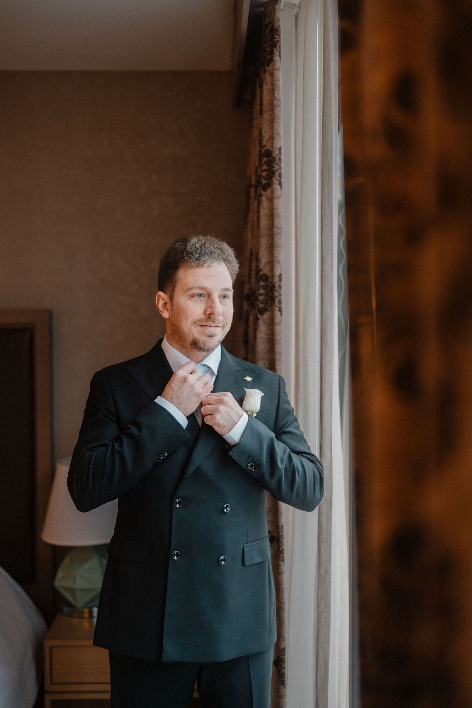 Dapper groom in a dark double-breasted suit adjusts his tie near a window with natural light.