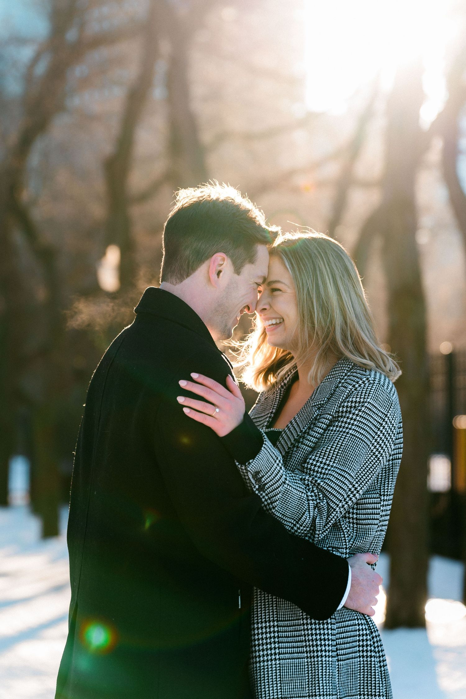 Smiling blonde woman in plaid coat with partner in sunny snowy park setting.