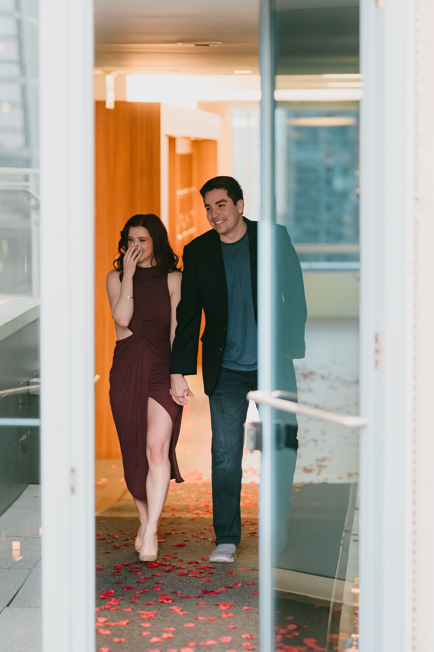 Couple holding hands walking through a doorway scattered with red rose petals in a warmly lit corridor.