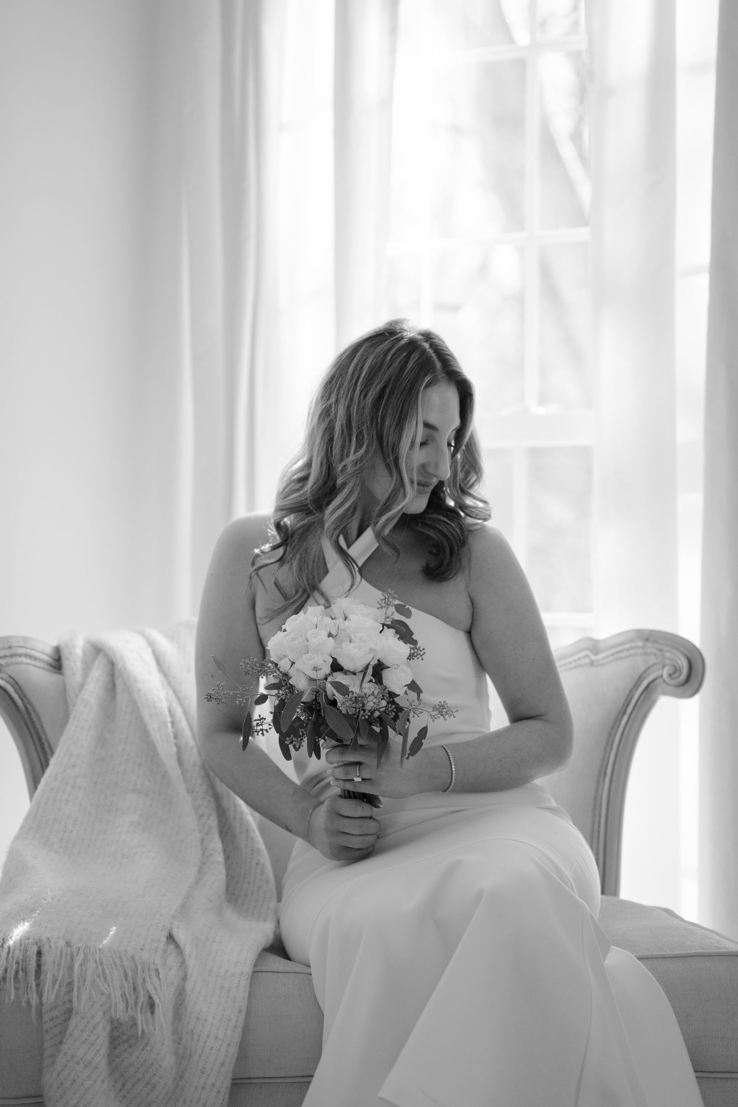 Black and white photo of bride in strapless gown sitting on sofa holding white bouquet.
