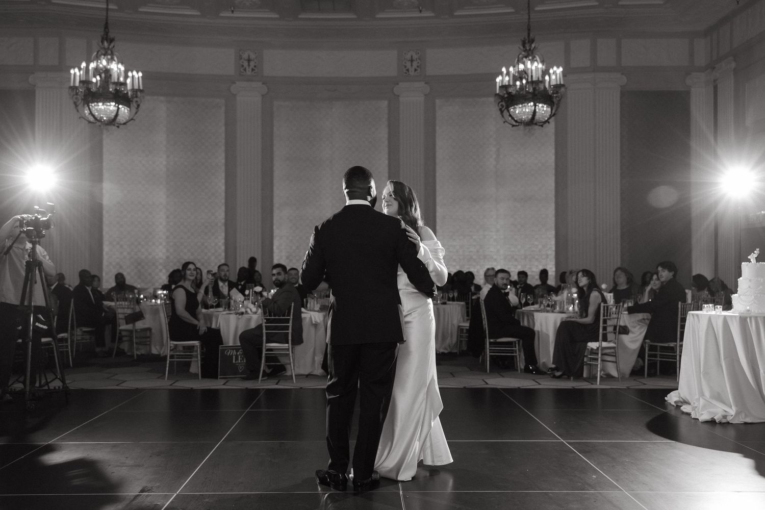 Black and white photo of a couple sharing their first dance at a wedding reception in an elegant ballroom.