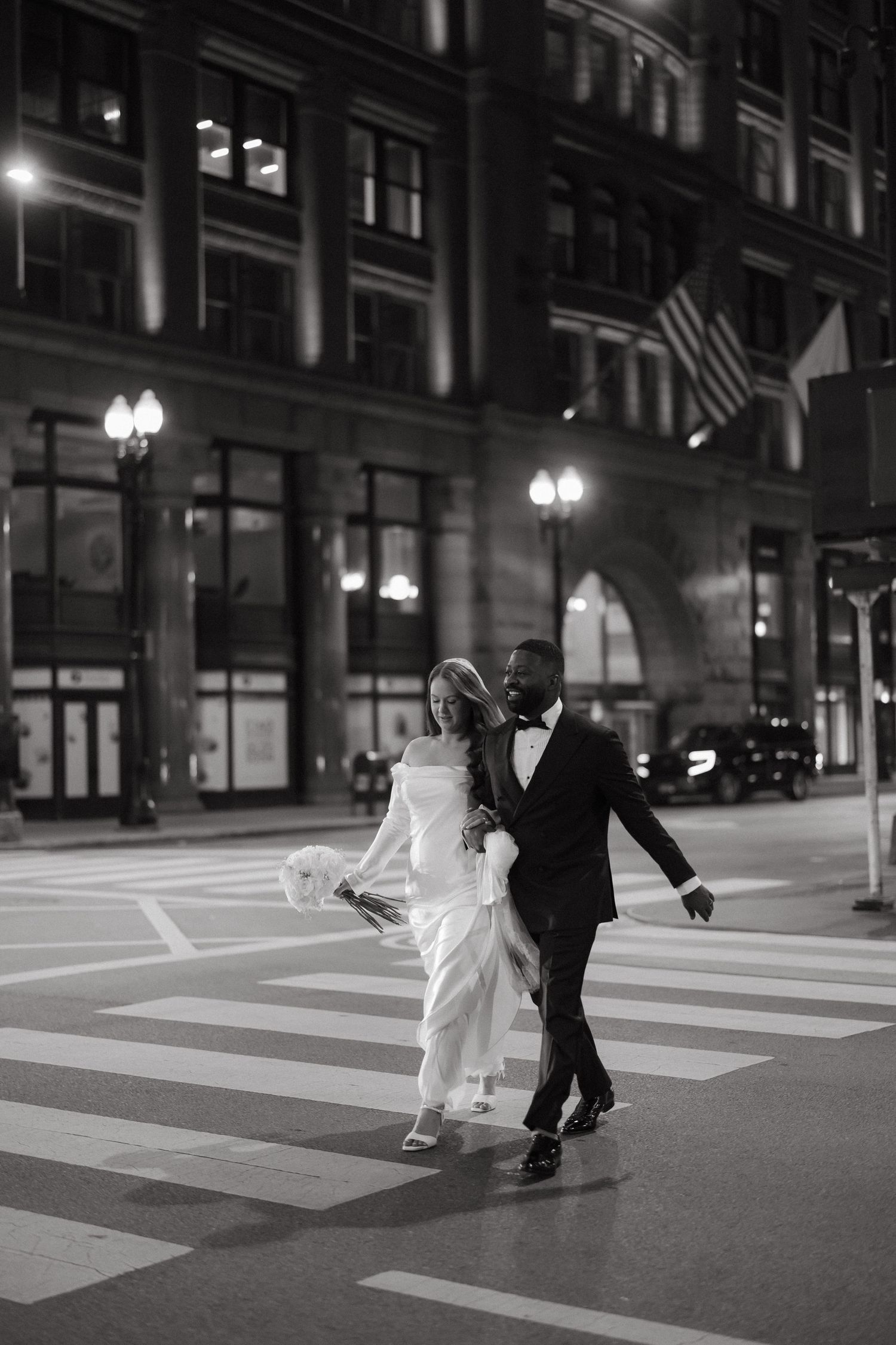 Black and white photo of a bride and groom crossing a city street at night, with grand buildings and street lights behind them.