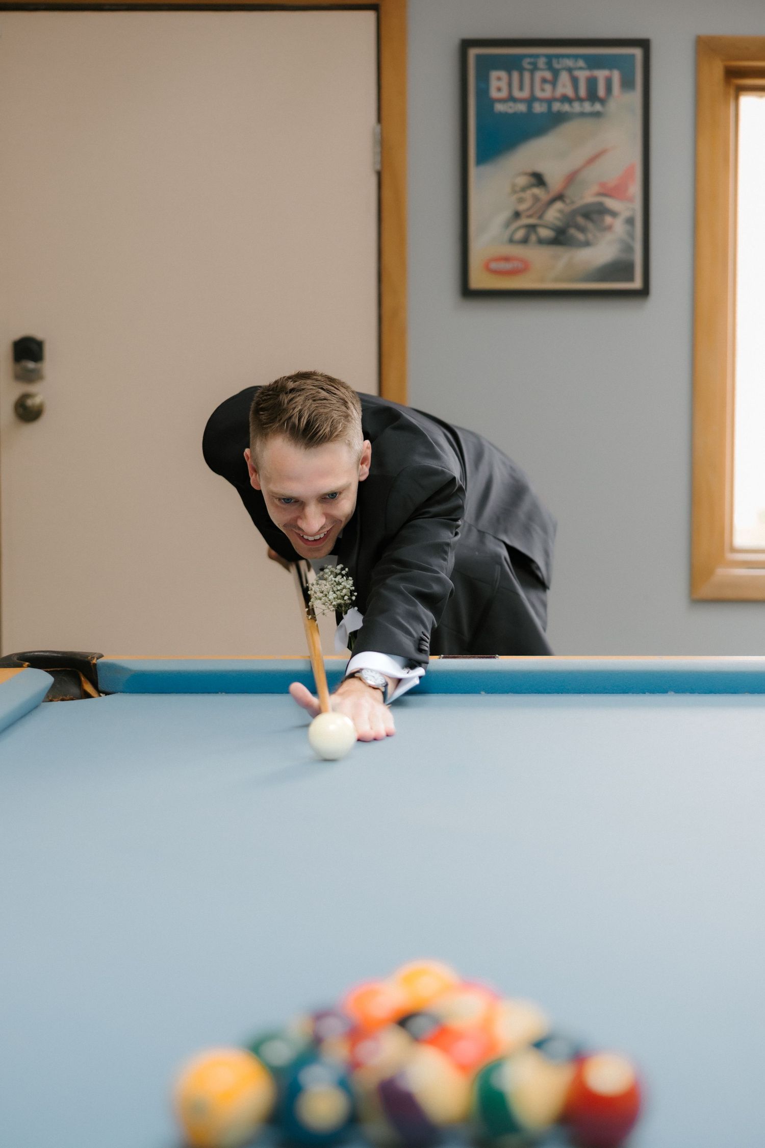 Groom in tuxedo smiling while playing pool billiards during wedding day preparations.
