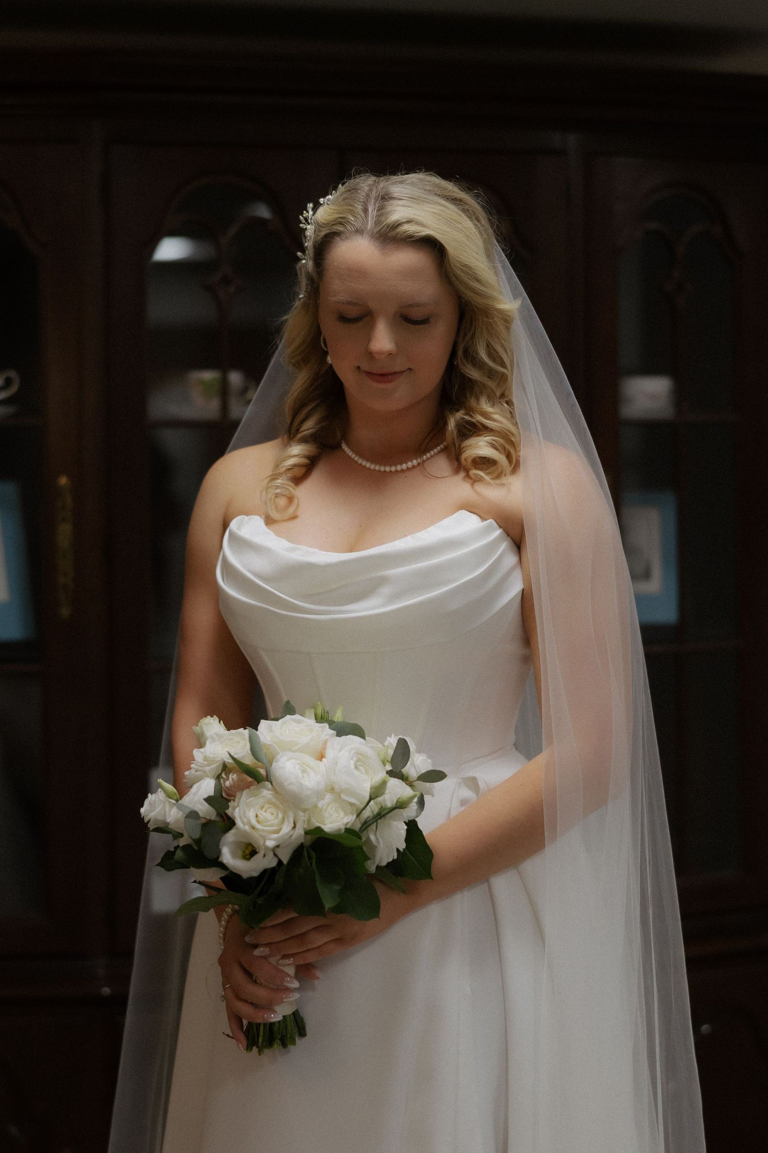 Elegant bride in strapless white gown holding bouquet of white roses, wearing veil and pearl necklace.