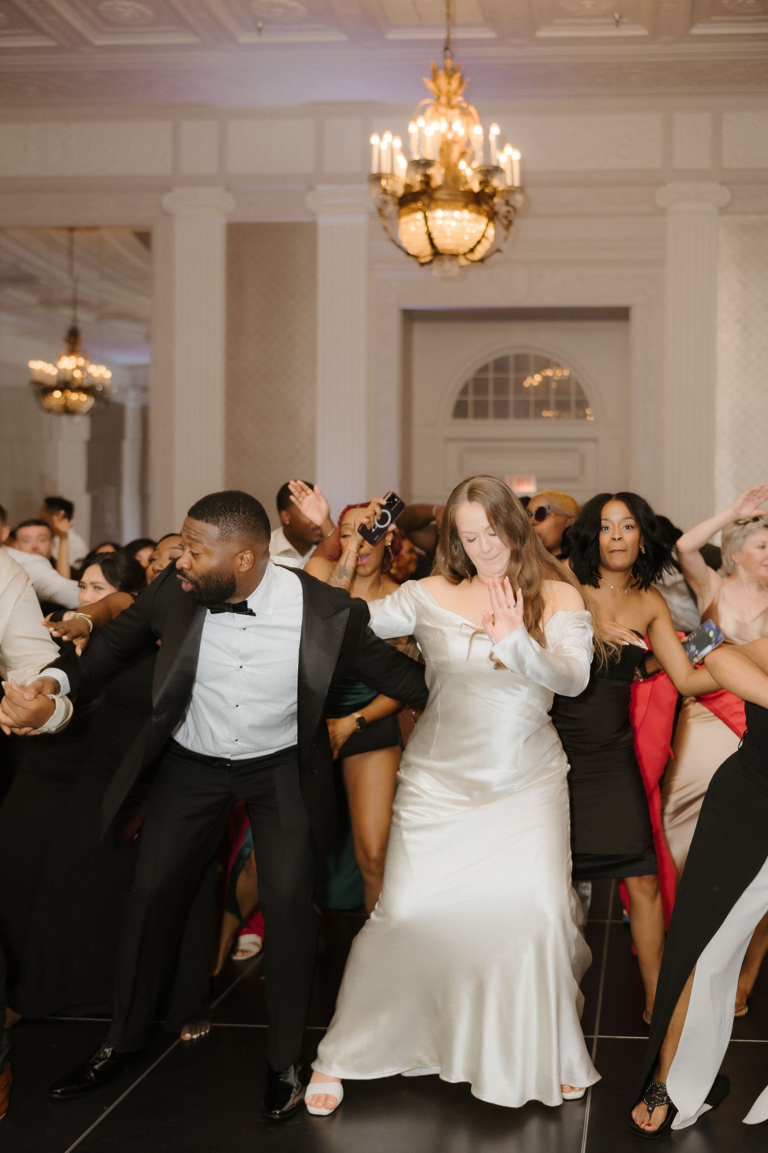 Bride and groom dancing joyfully with guests at elegant wedding reception under golden chandelier.