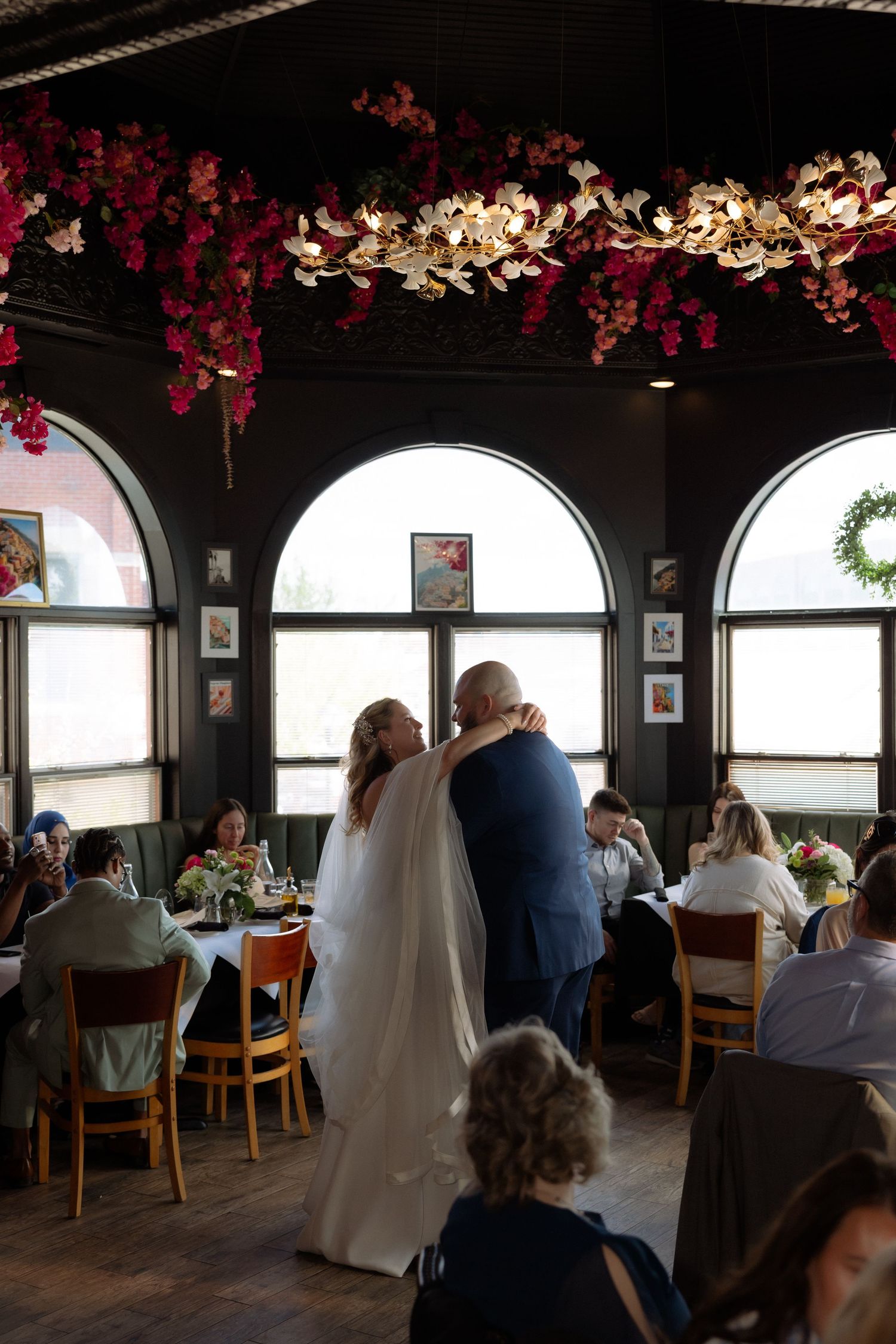Bride and groom share first dance in elegant restaurant venue adorned with red floral ceiling garlands and arched windows.