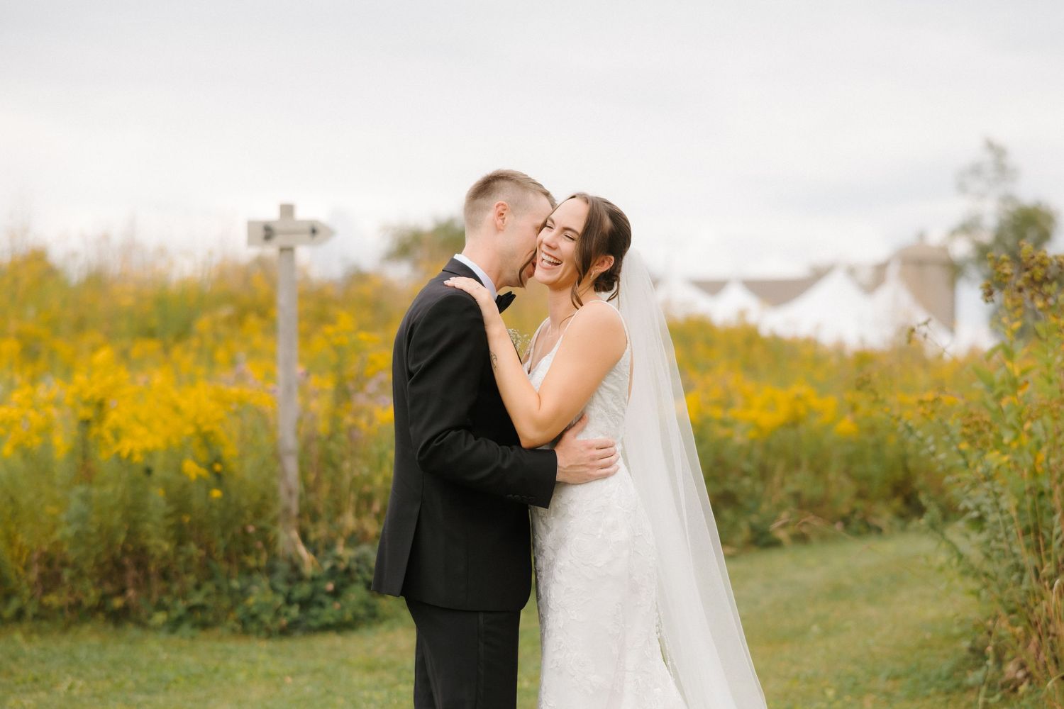Groom tenderly kissing bride on cheek in yellow wildflower field with cross visible in background.