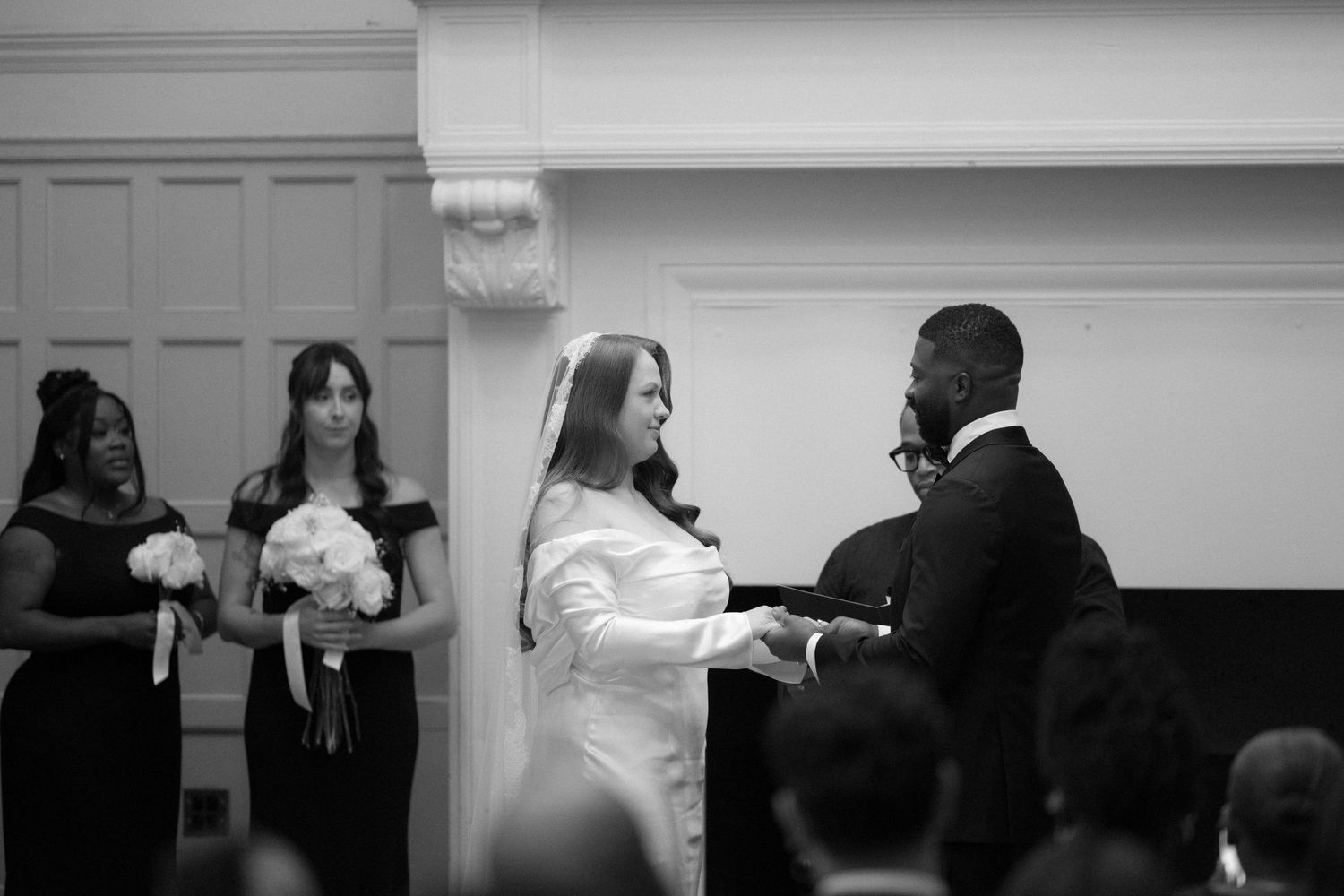 Black and white wedding ceremony photo showing bride in white gown exchanging vows with groom, bridesmaids in black dresses behind.