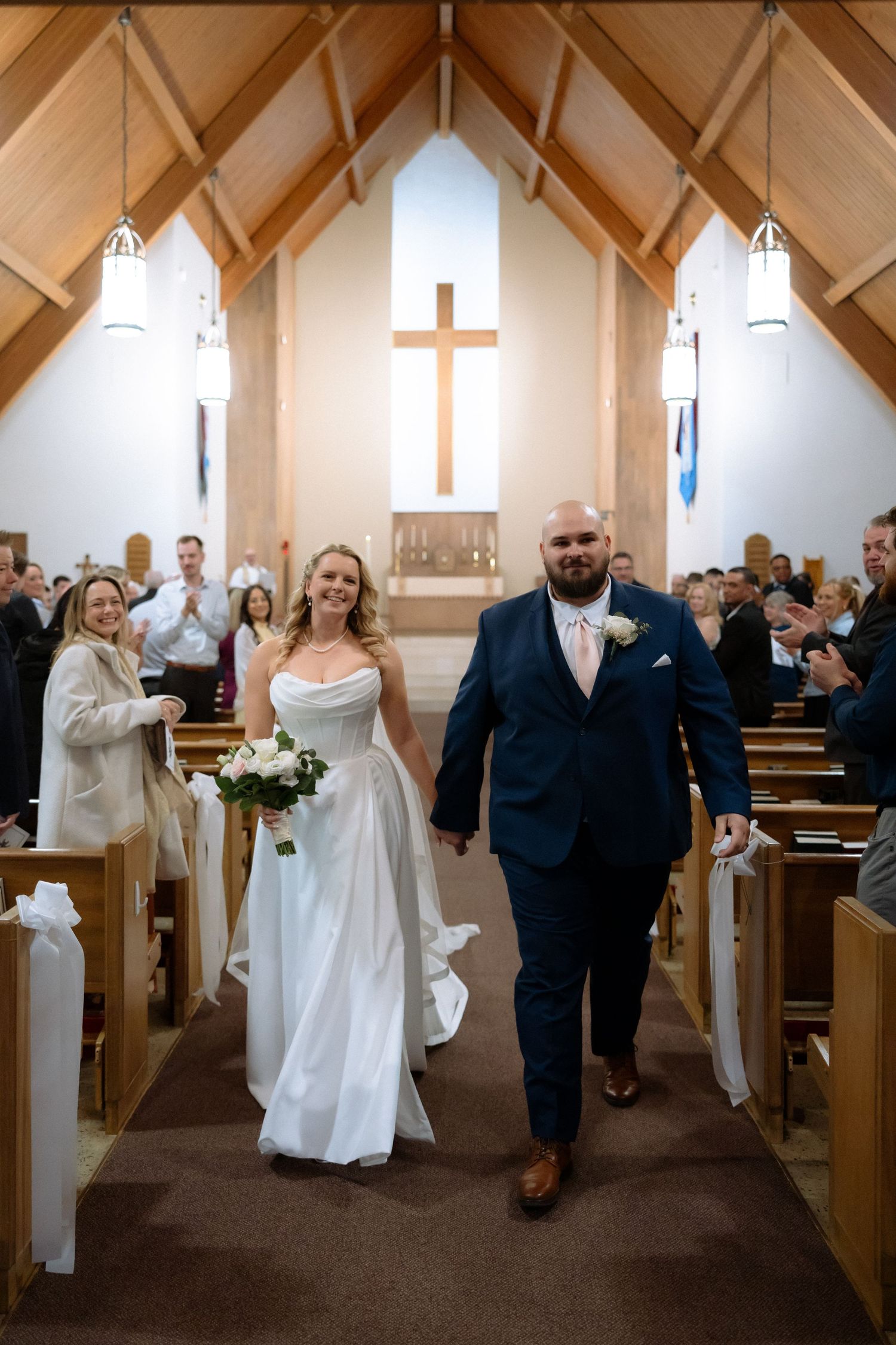 Bride and groom walk down church aisle after wedding ceremony, bride holding white bouquet, cross visible at altar.