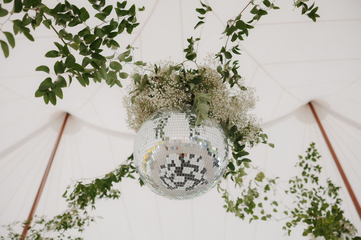 Disco ball decorated with greenery and white flowers hanging from a white wedding tent ceiling.