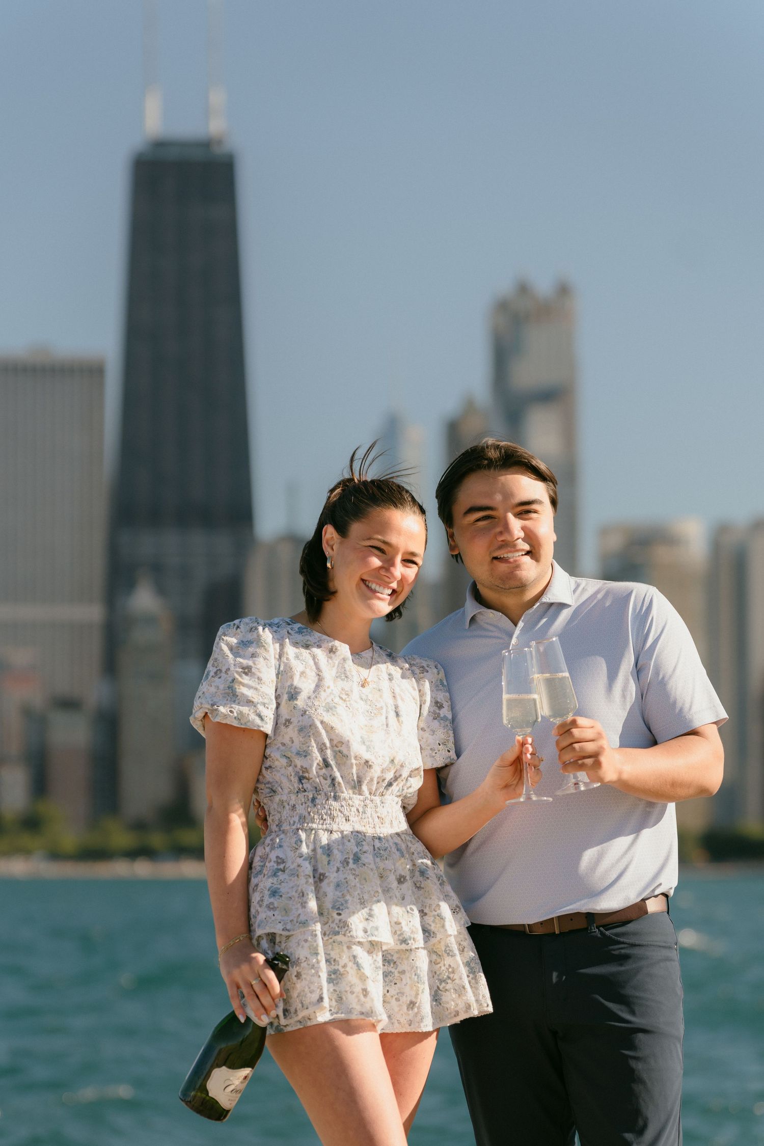Smiling engaged couple toasting champagne glasses with Chicago skyline in the background.