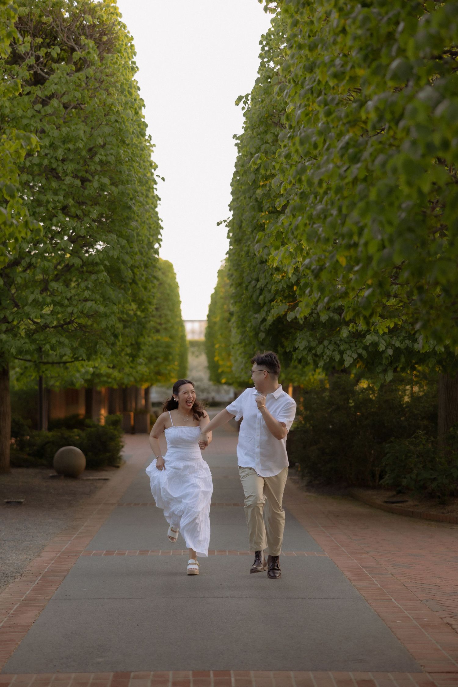 Couple in wedding attire joyfully running hand-in-hand down a tree-lined pathway during golden hour.