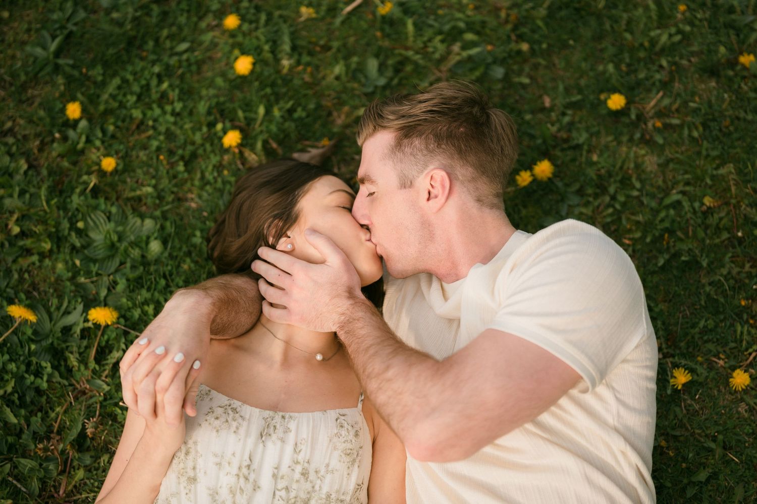 Couple lies in grass with yellow wildflowers as man kisses woman on the cheek.