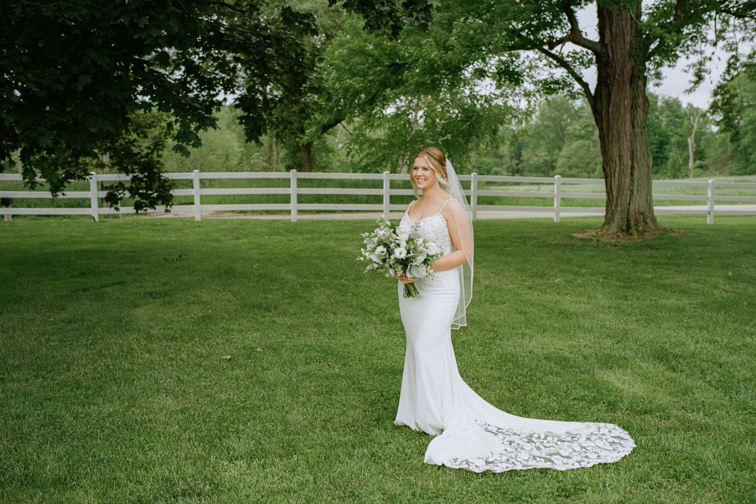 Bride in strapless white mermaid gown with lace train holding bouquet on green lawn near white fence and trees.