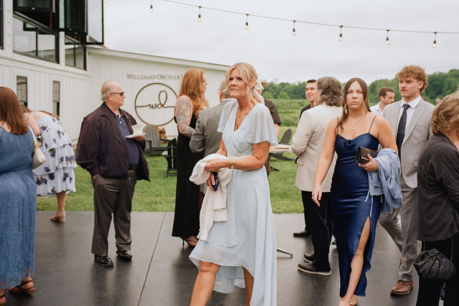 Guests mingle outdoors at a wedding reception, woman in light blue midi dress holding white clutch.