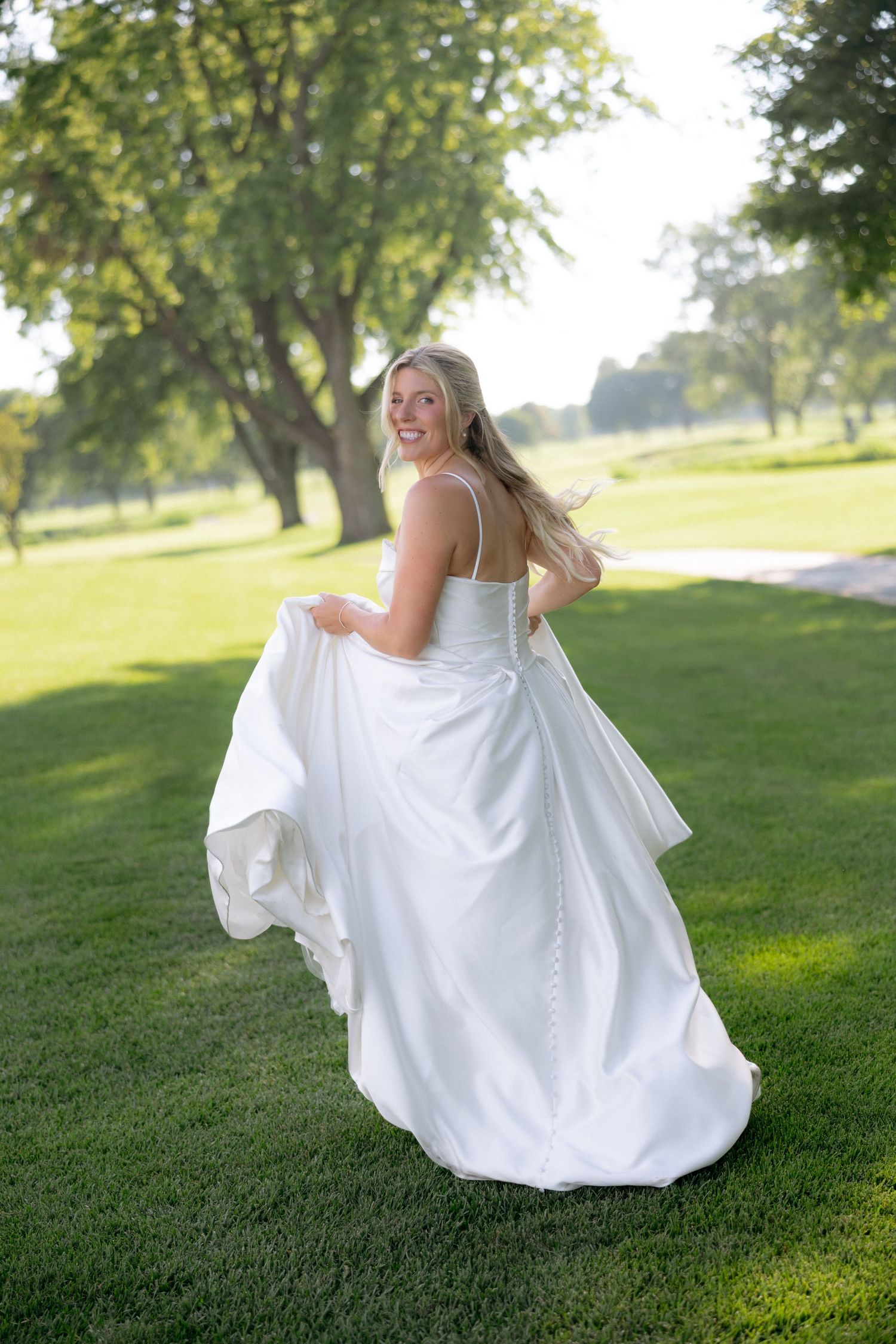 Radiant bride in strapless white gown twirls on lush green lawn during sunny outdoor wedding photo session.