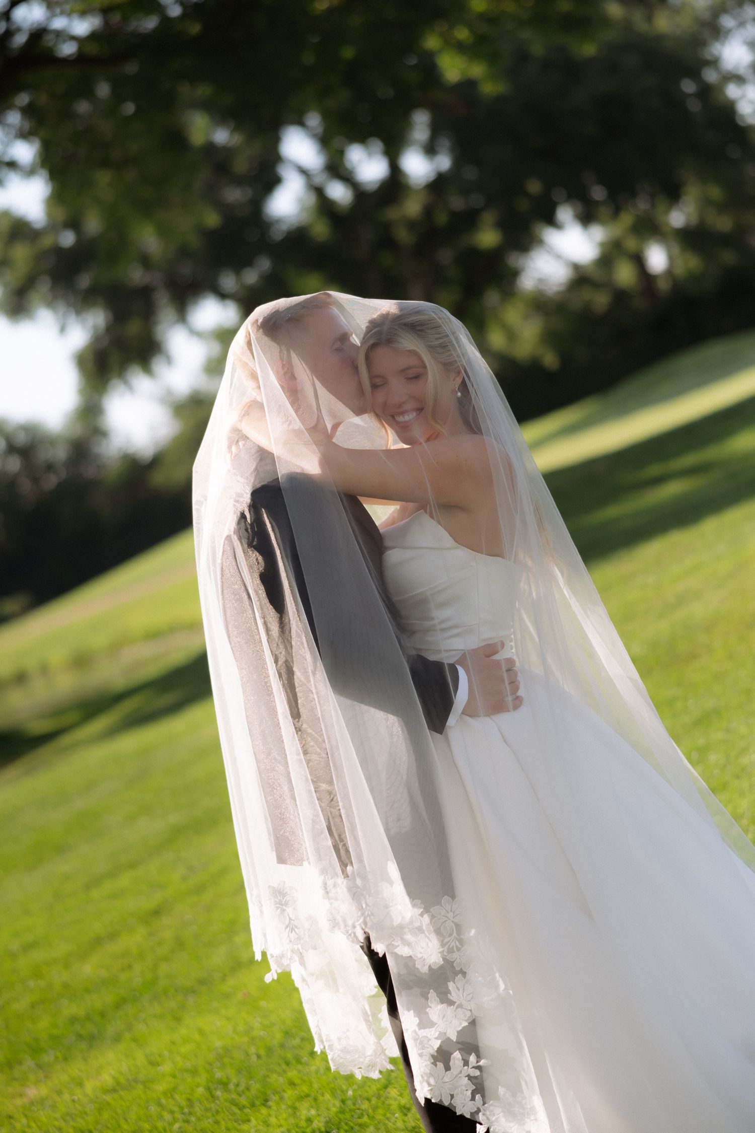 Groom kisses bride under flowing lace veil in sunlit green park during romantic outdoor wedding portrait.