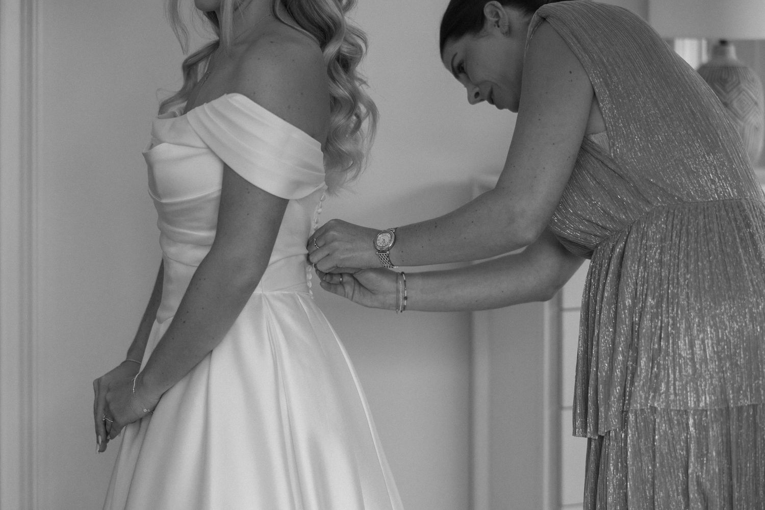 Black and white photo of a woman helping a bride button her white off-shoulder wedding dress.