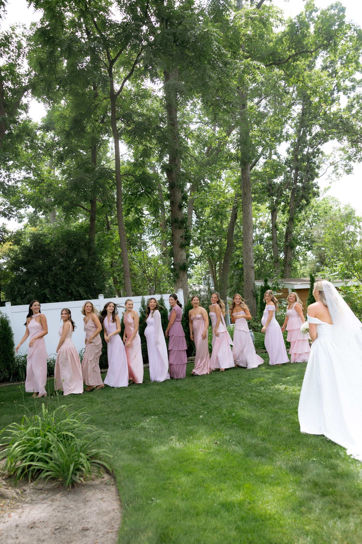 Bride in white gown facing row of bridesmaids in pink dresses for first look outdoors.