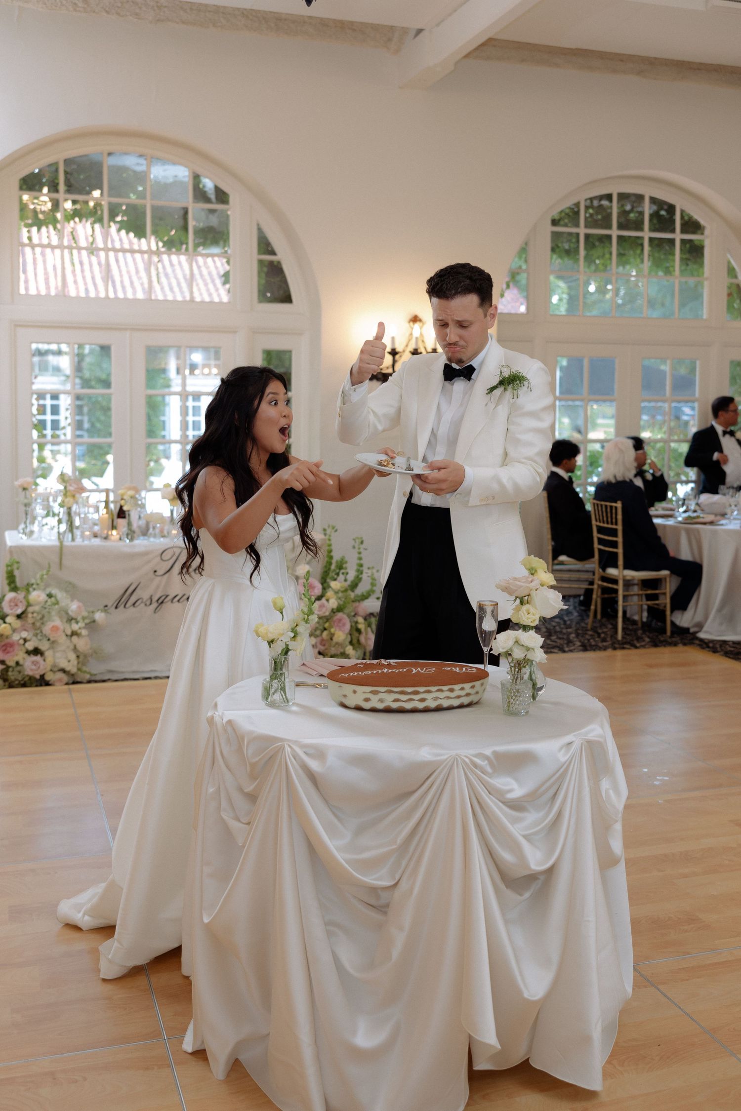Bride and groom playfully serve wedding pie at their elegant white-decorated reception venue.