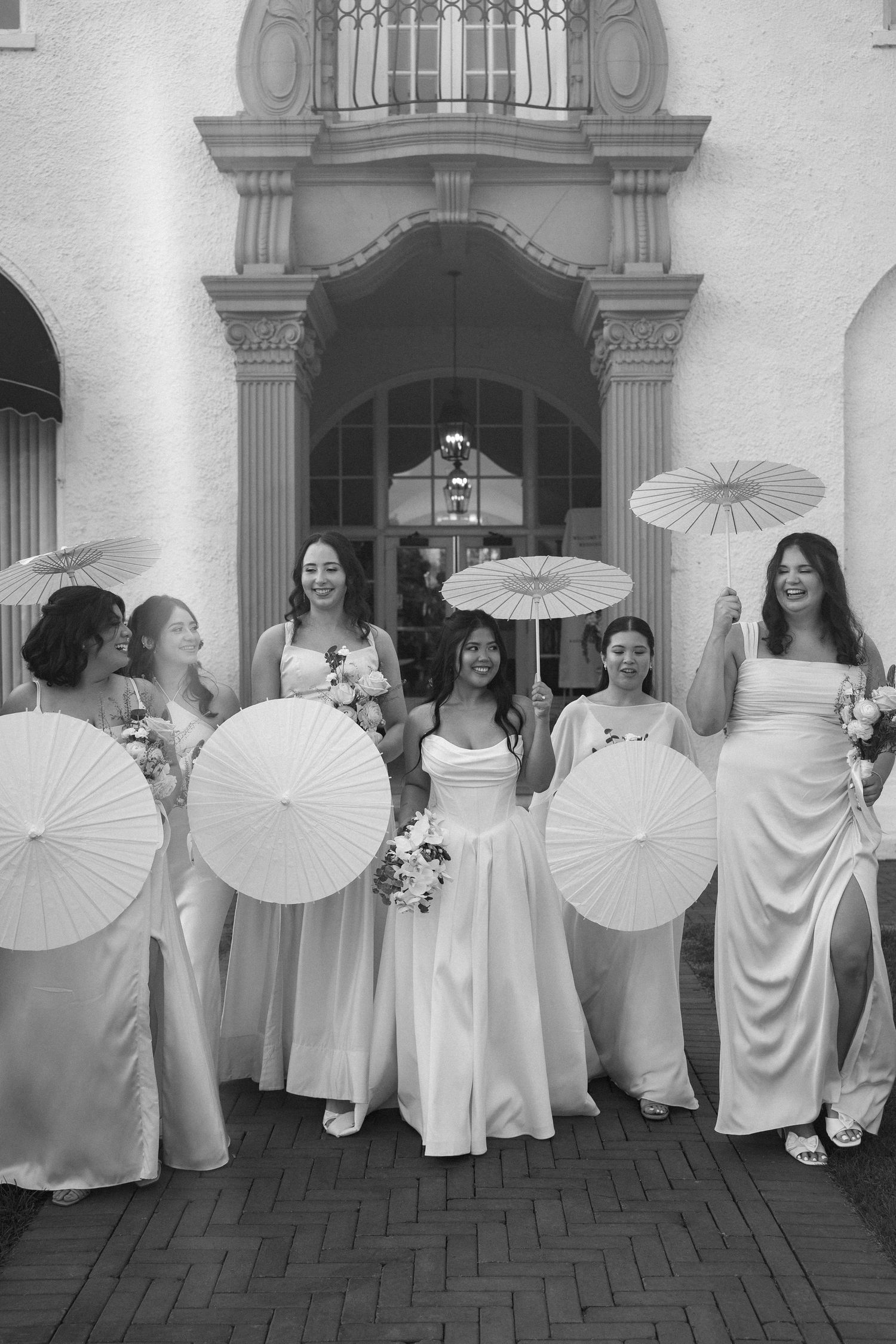 Smiling bride and bridesmaids in white dresses carry parasols outside ornate building in black and white photo.