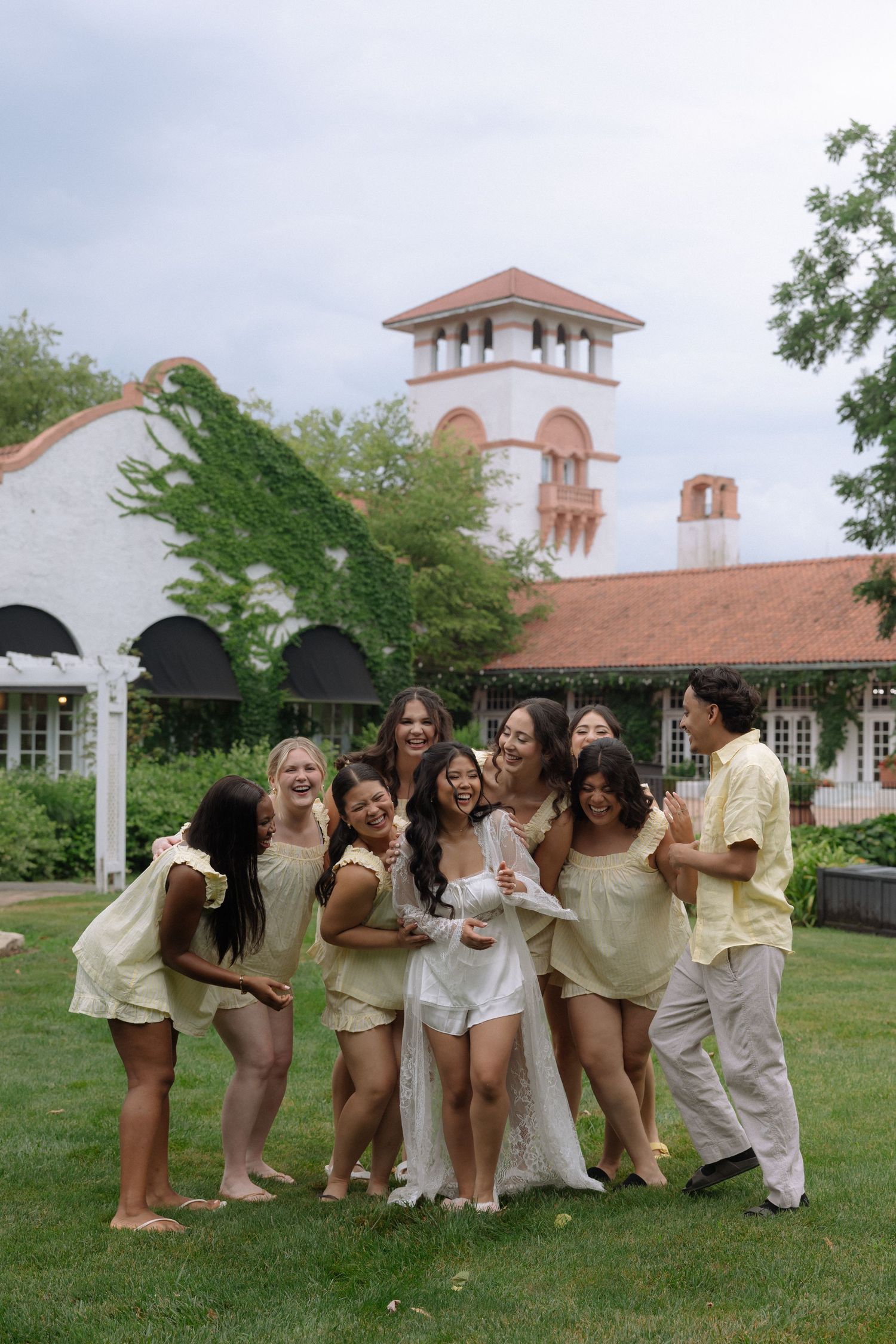 Bride in white lace gown laughing with bridesmaids in yellow outfits on green lawn outside elegant venue.
