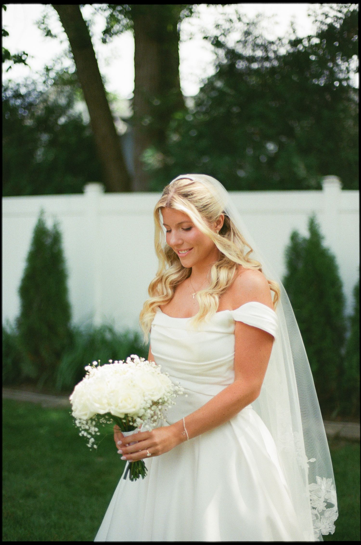 Bride in off-shoulder white gown holds white floral bouquet outdoors, smiling softly with veil and blonde hair.