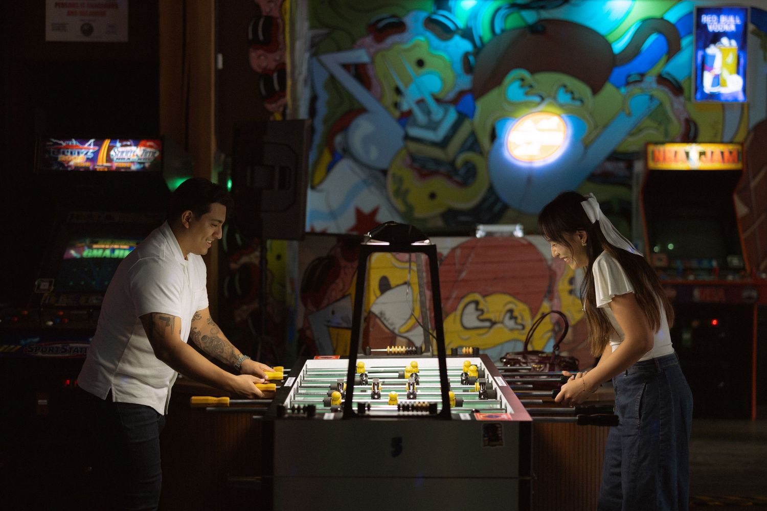Two people playing foosball in a dimly lit arcade with colorful game machines in the background.