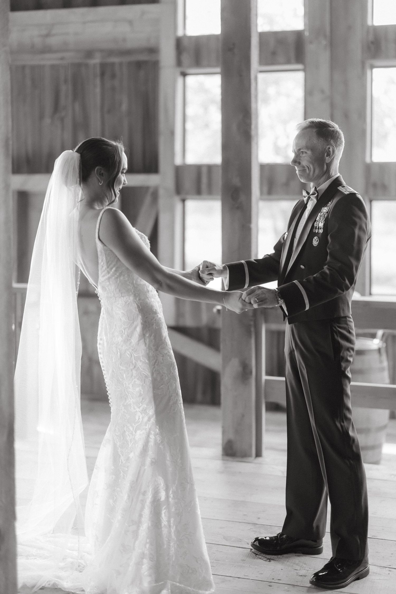 Black and white photo of a bride in lace gown holding hands with a military officer during a wedding first look.