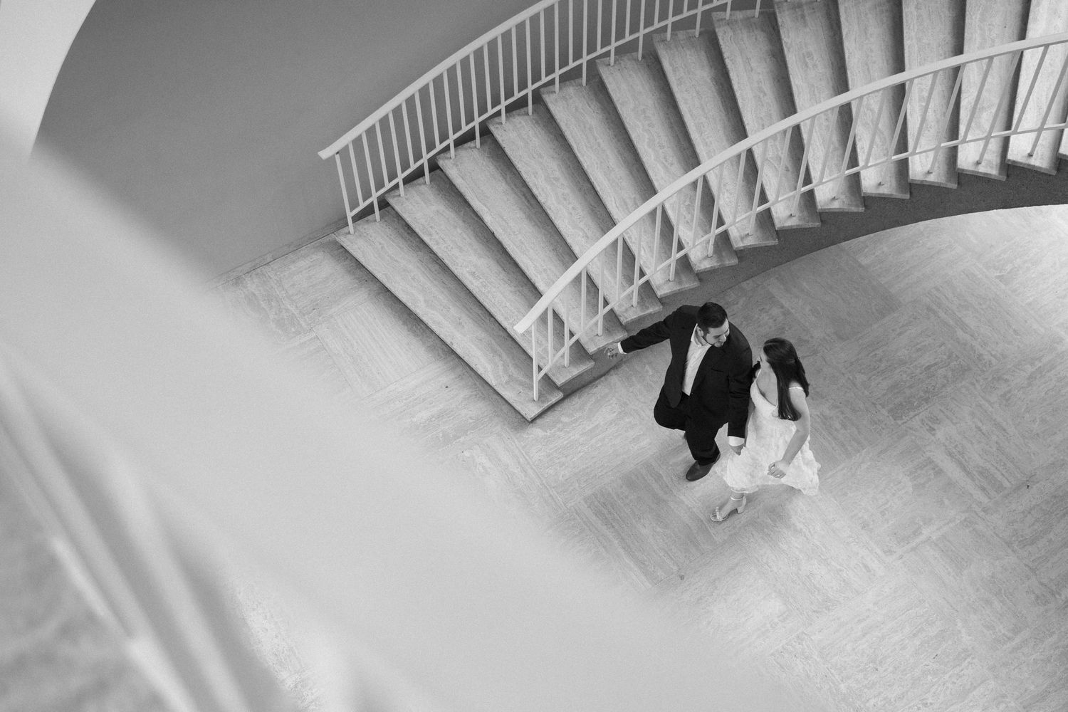 Black and white aerial photo of a bride and groom dancing beneath a curved staircase.