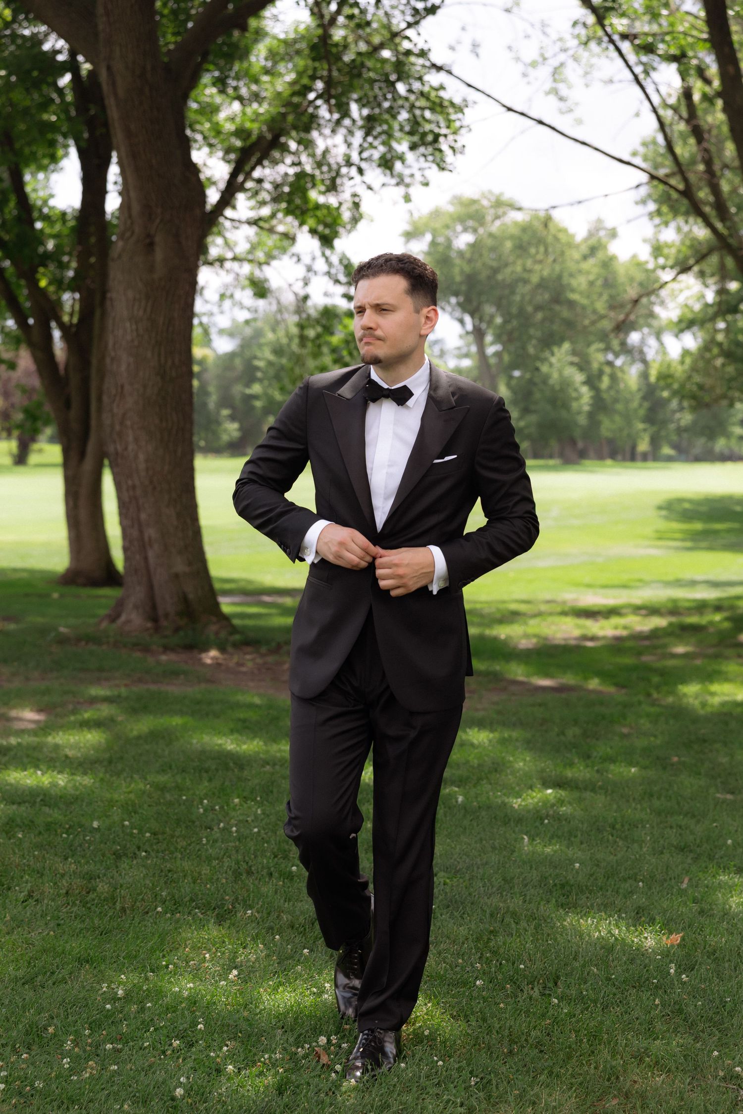 Handsome groom in black tuxedo and bow tie buttons jacket while standing in green park setting.