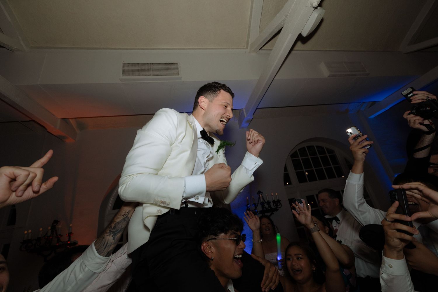 Groom in white tuxedo is lifted on shoulders by cheering guests during lively wedding reception celebration.